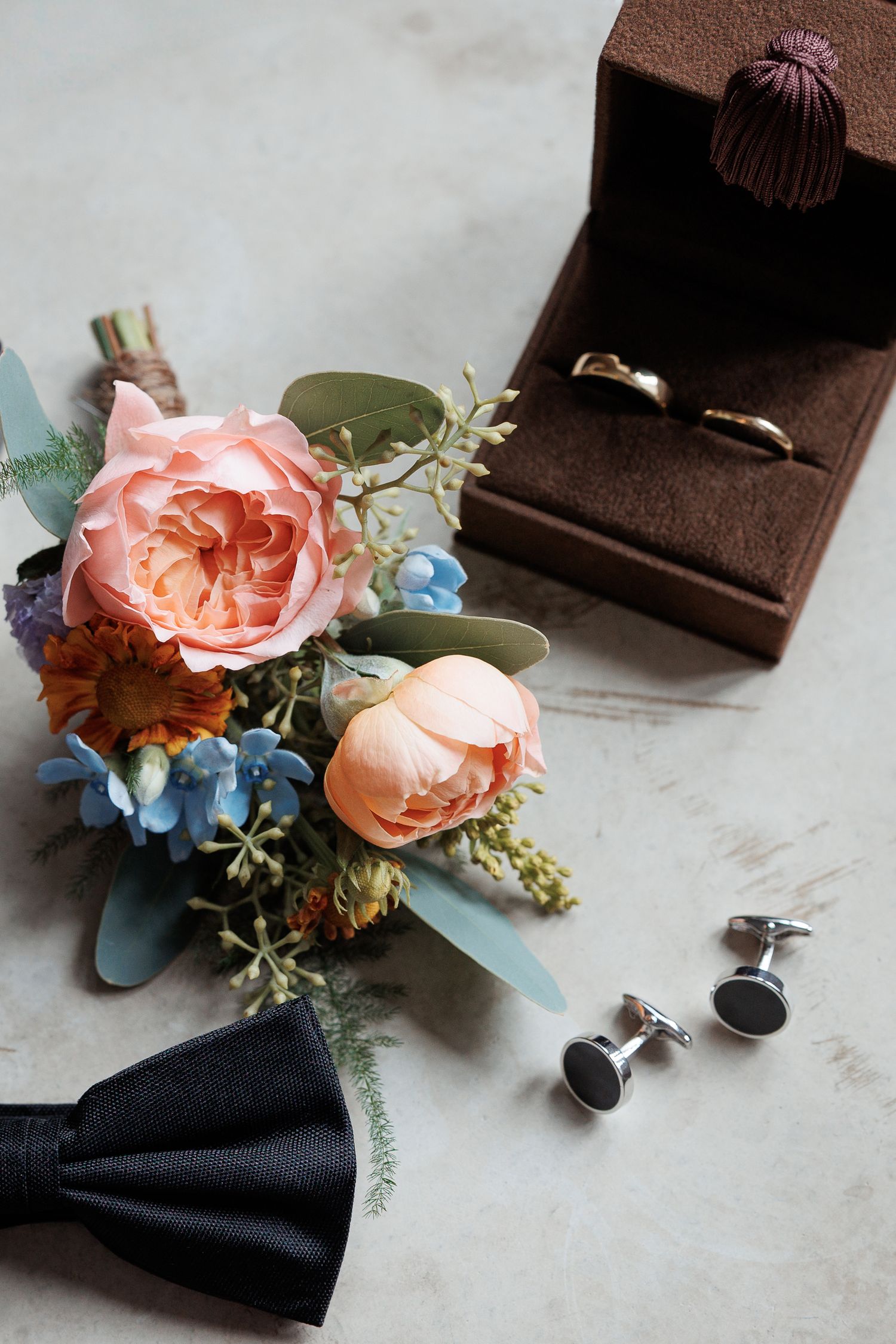 Wedding accessories with pink roses, blue hydrangea boutonnieres, cufflinks and black tie arranged on marble surface.