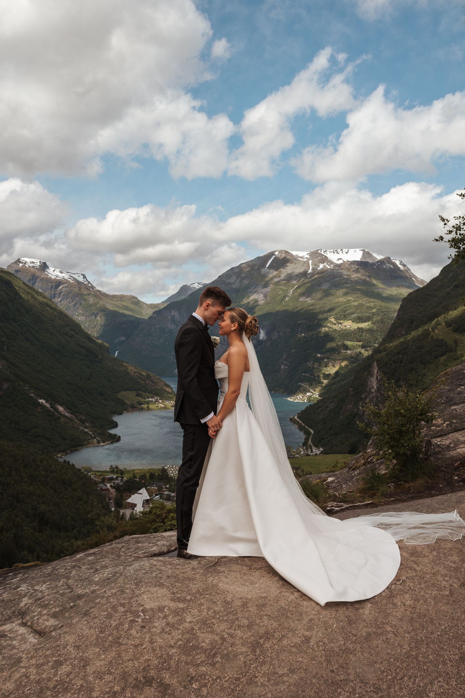 A wedding couple stands on a mountain overlook with snow-capped peaks and a serene lake valley in the background.