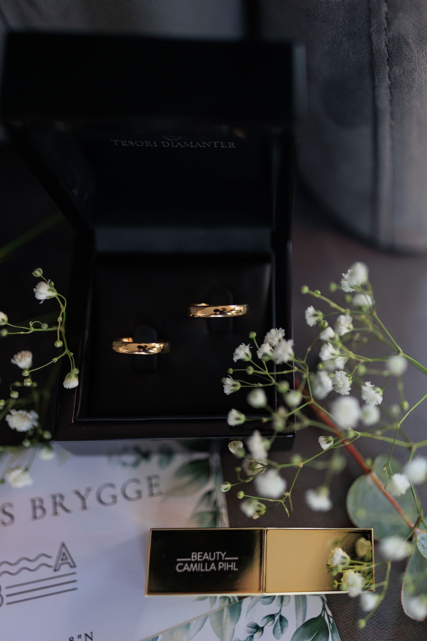 Two gold wedding rings in a black box surrounded by delicate baby's breath flowers and branding materials.