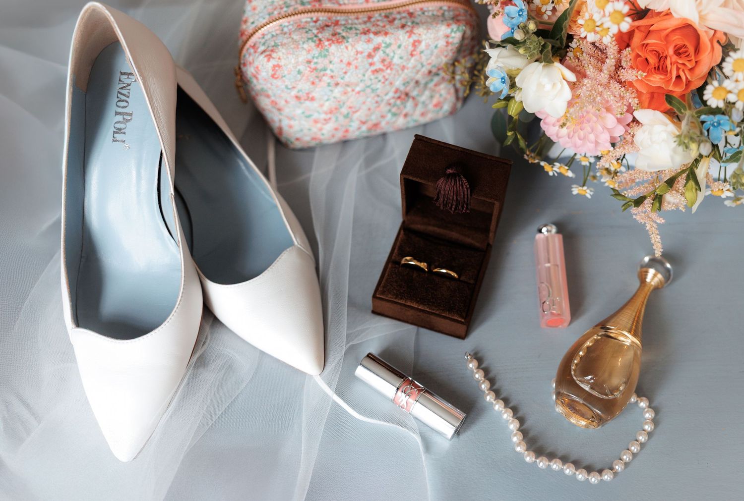 Elegant flat lay of pink and white bridal accessories including jewelry, veil, shoes, and florals on light surface.
