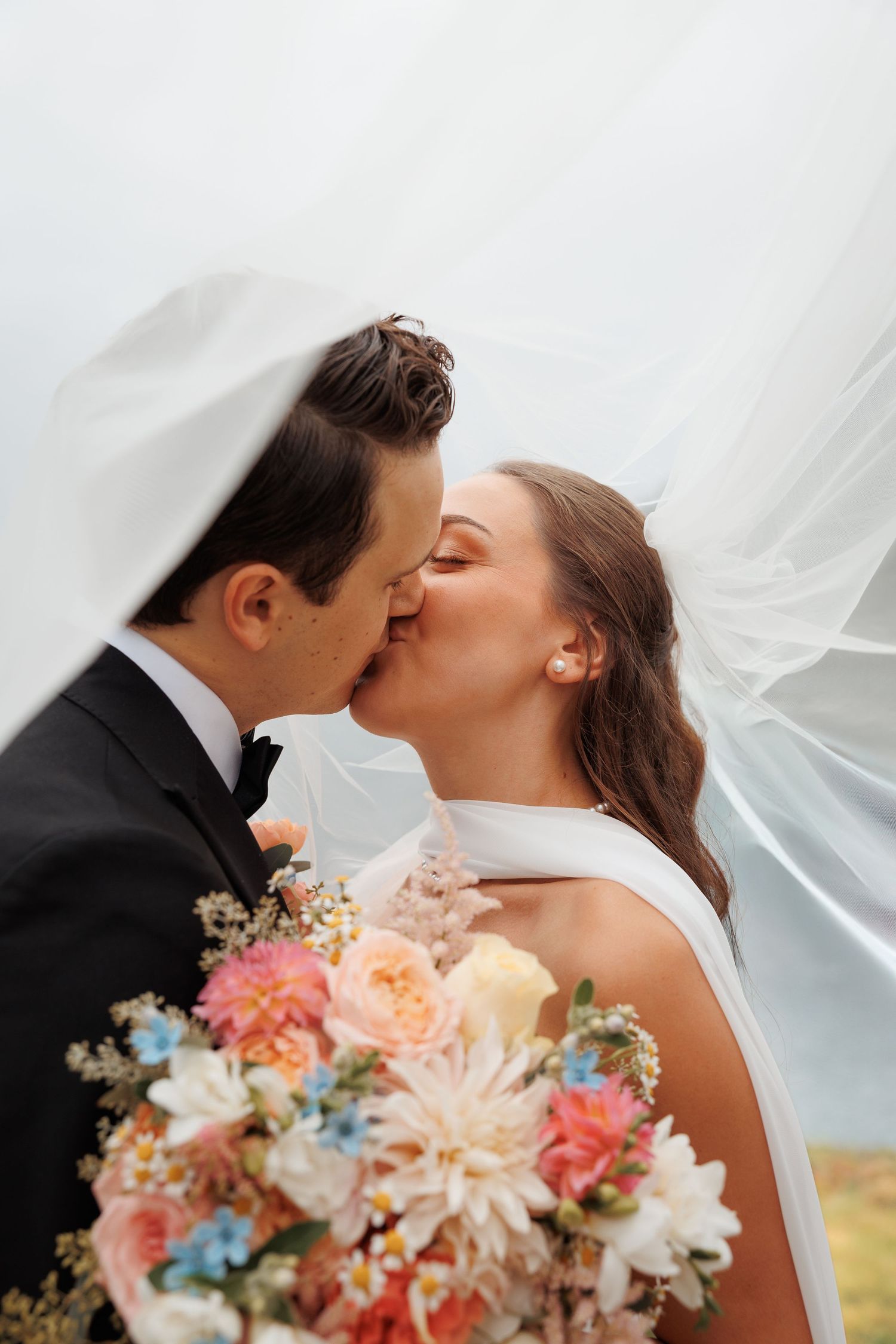 Romantic wedding portrait with bride holding peach and blue floral bouquet as a veil flows in the breeze.