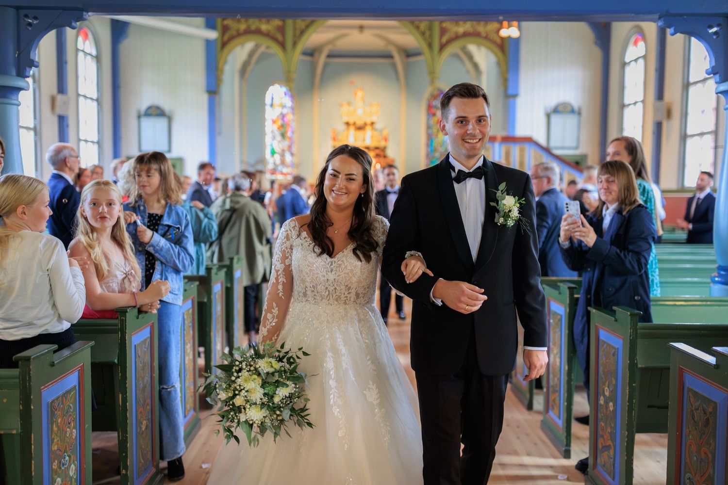 A couple walks down the aisle of a beautifully decorated church surrounded by wedding guests.