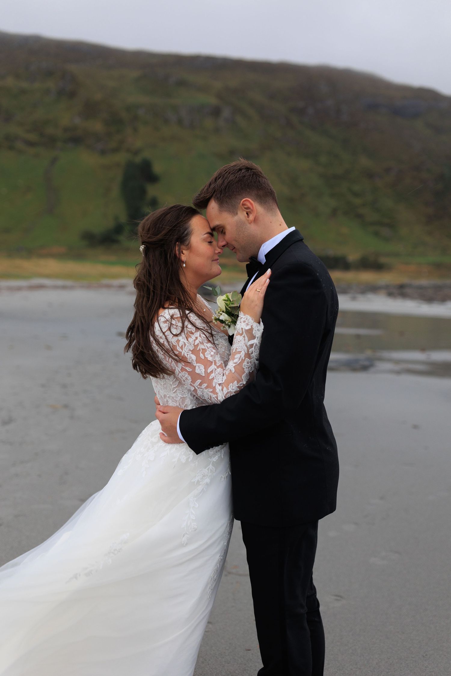 Bride in lace sleeve gown and groom share tender moment on beach with rolling hills in background.