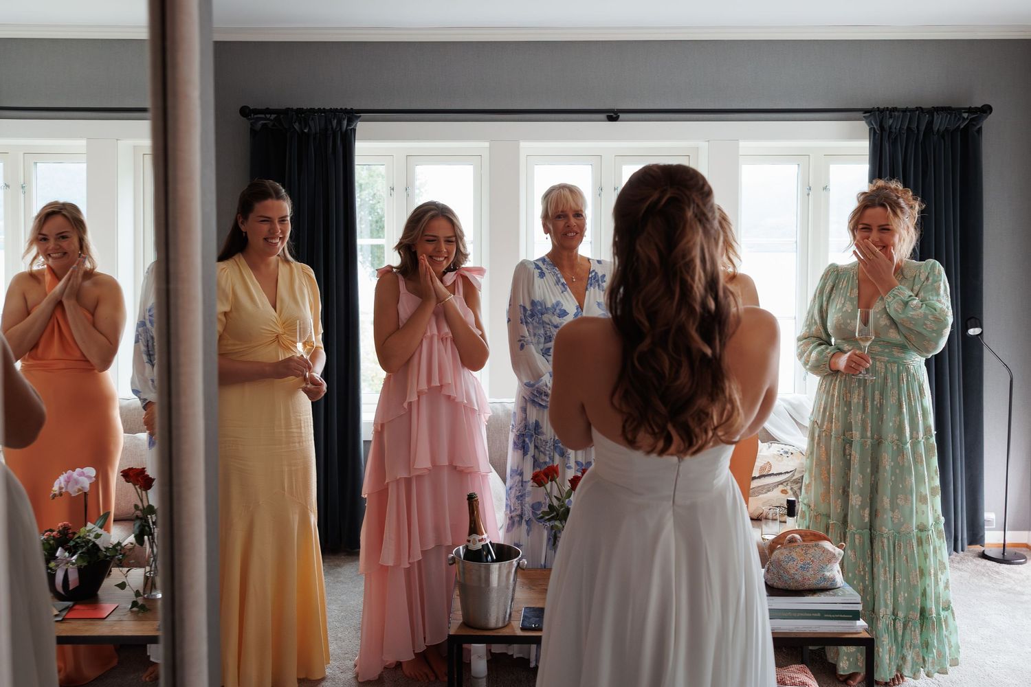 Several people in pastel colored dresses stand in a bright room together near a window and dark curtains.