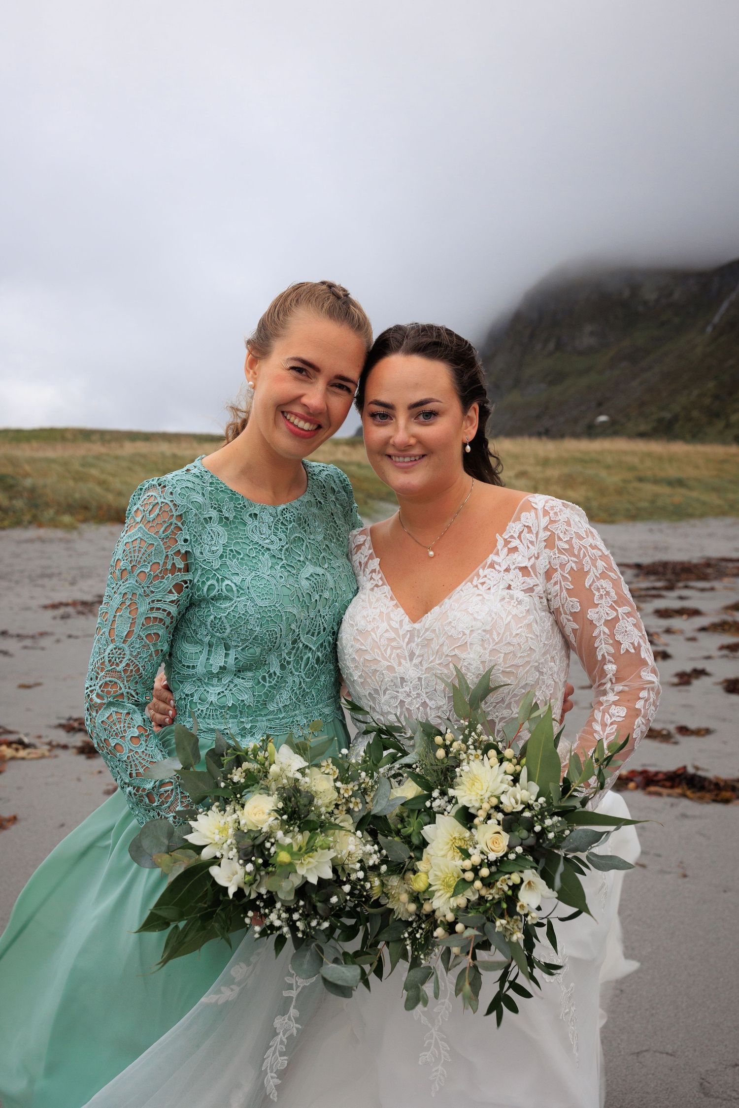 Wedding party members in mint green and white lace dresses pose with elegant bouquet on beach.