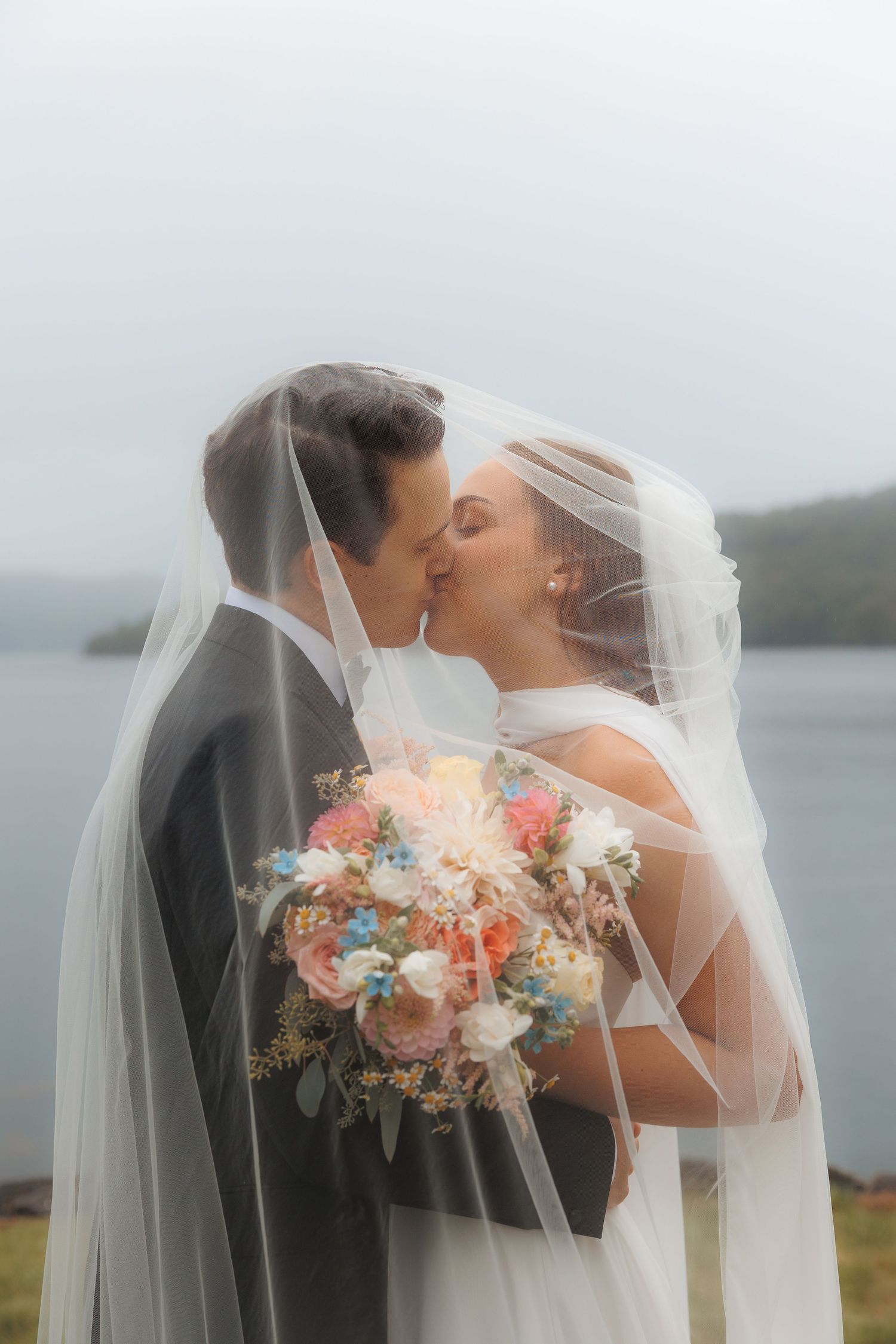 Romantic wedding couple embraces under a veil with a soft-focused floral bouquet in pastel colors.