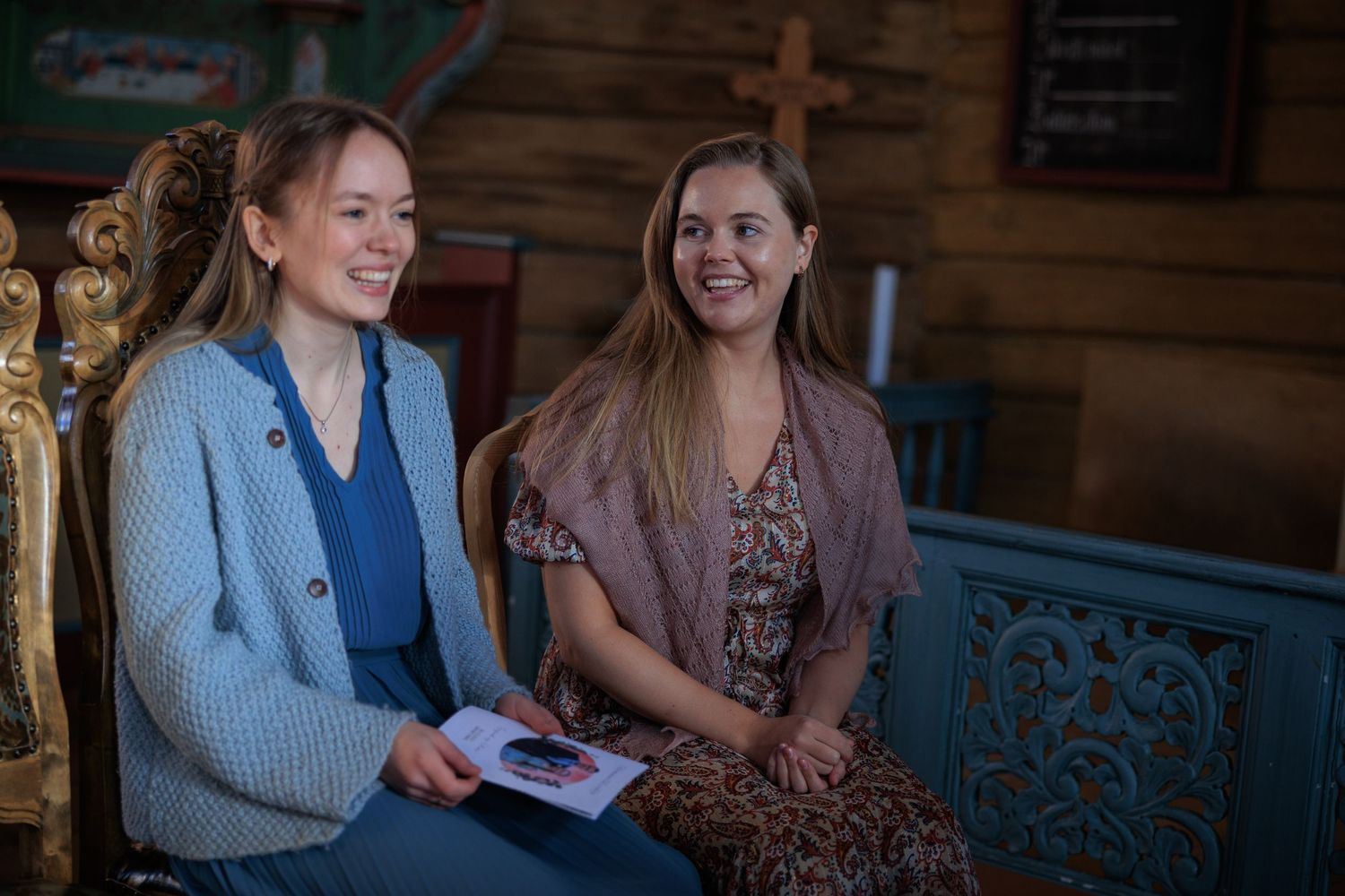 Two women chat while sitting on wooden bench in casual indoor setting.
