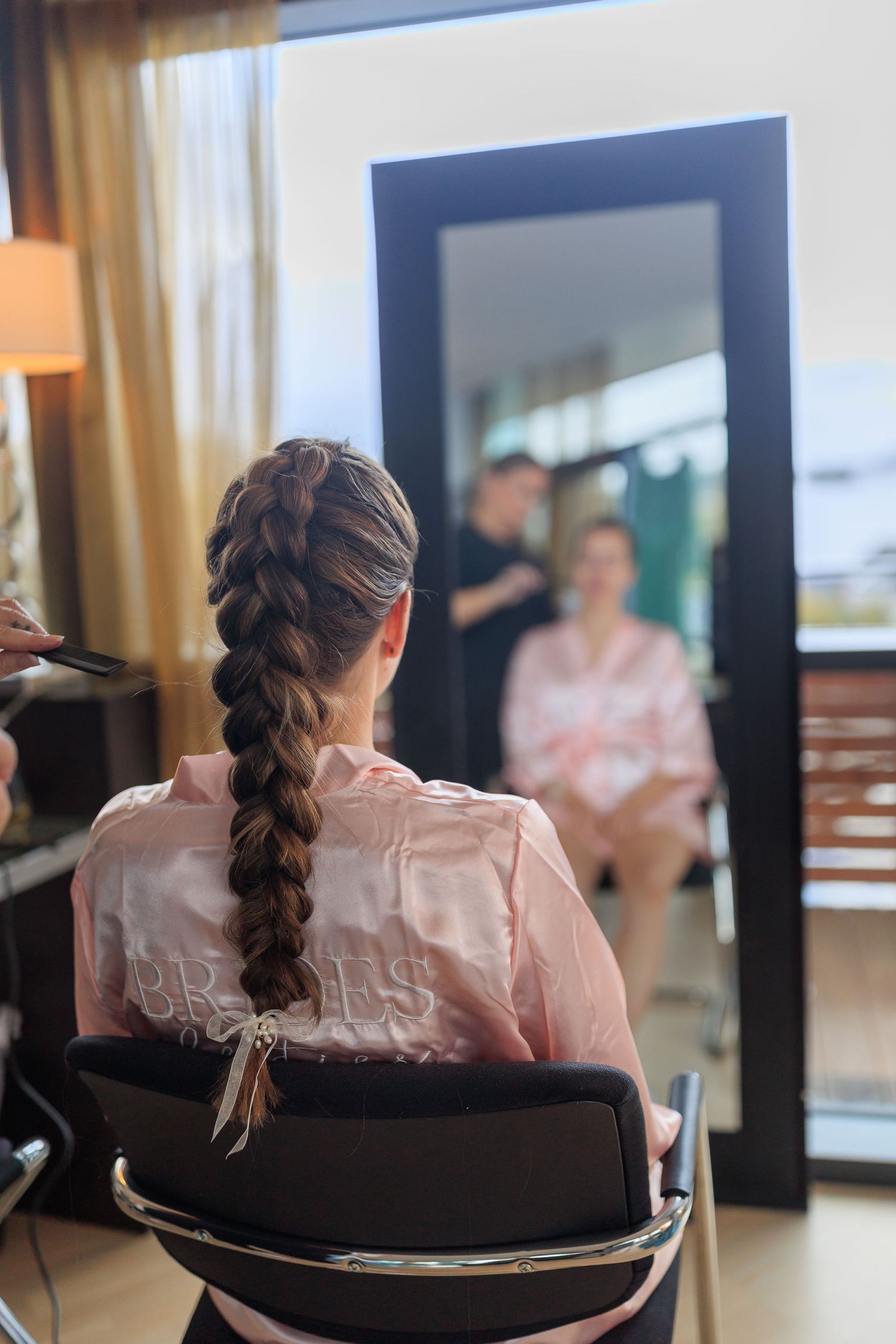 A person with a long braided hairstyle sits in a salon chair during wedding preparations.