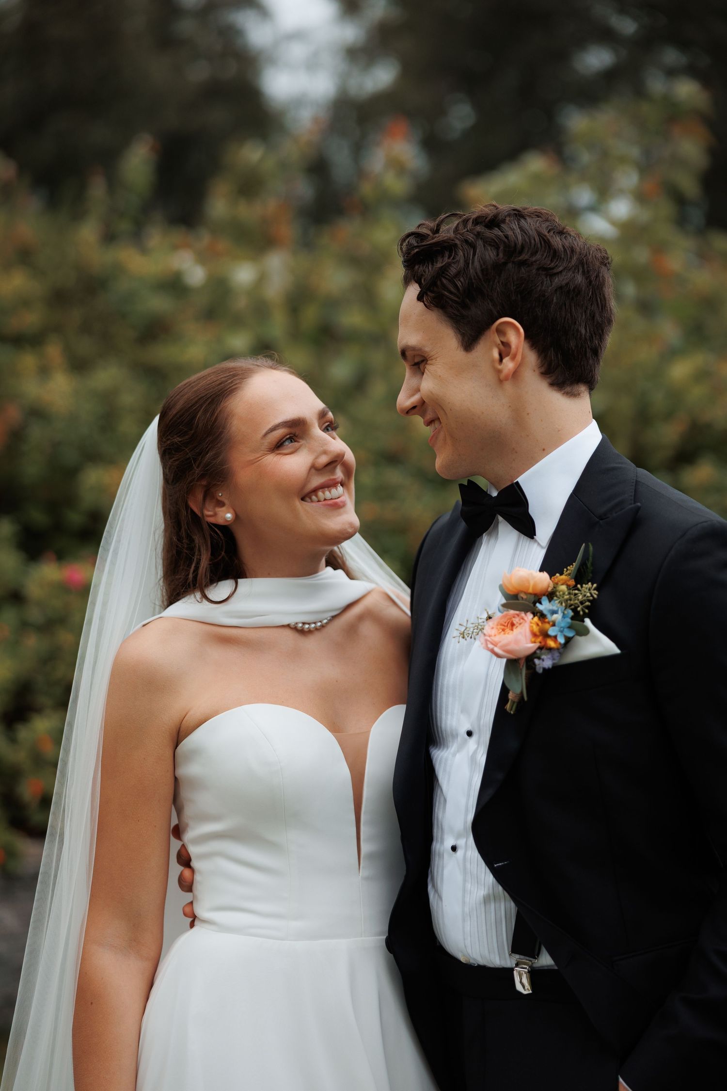 A happy couple shares a joyful moment on their wedding day, wearing a white gown and black tuxedo in an outdoor garden setting.