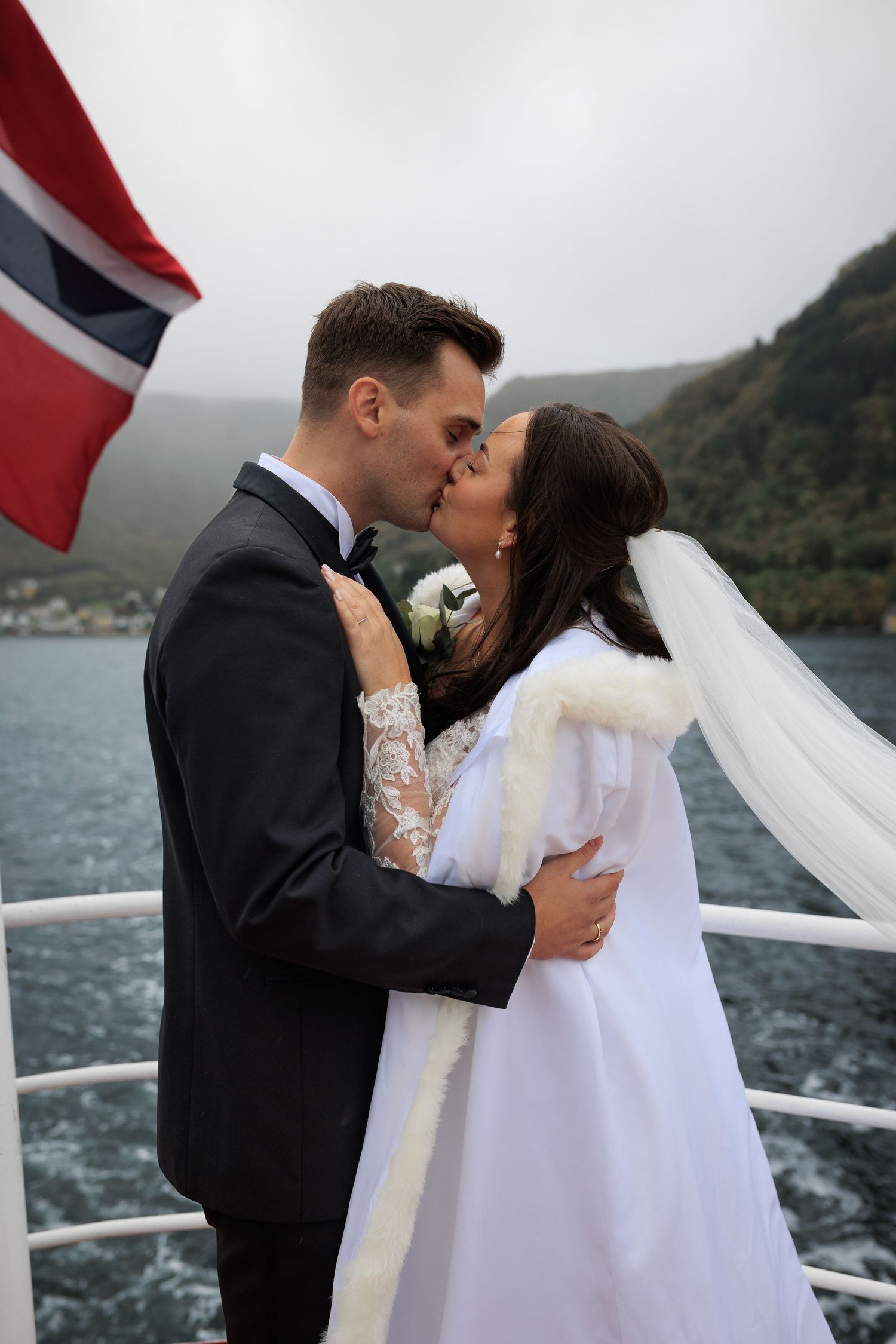 Couple shares romantic kiss on boat deck with Norwegian flag and mountainous fjord backdrop.