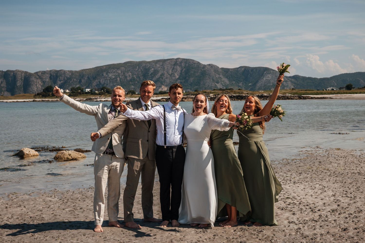 Wedding party celebrating on a beach with mountains in the background, bride in white and bridesmaids in green.