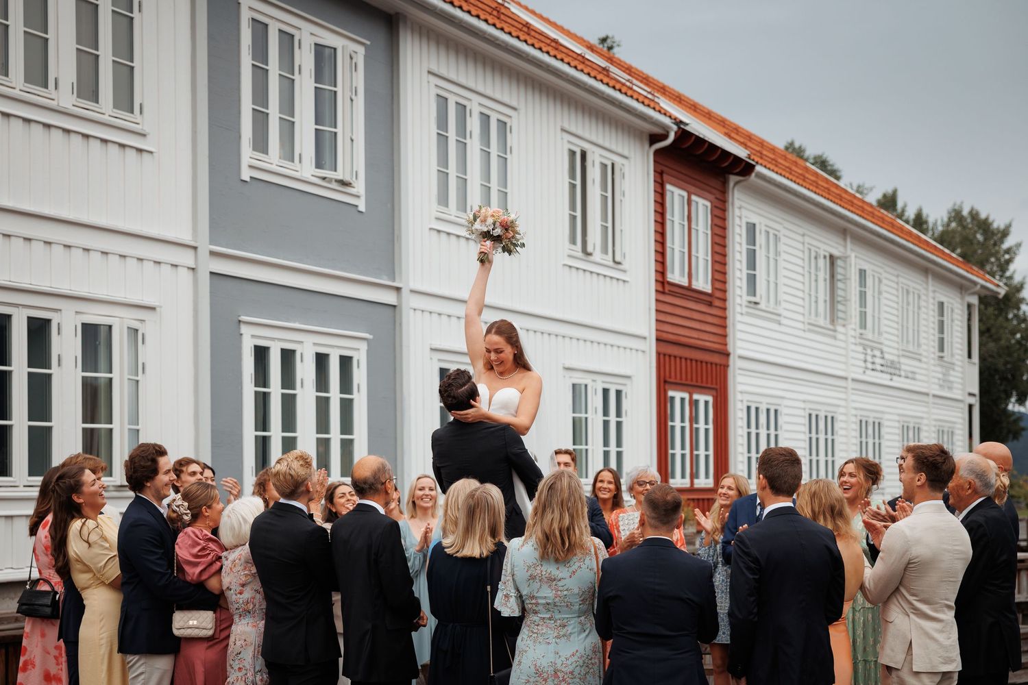 Wedding guests gather and socialize outside on a deck patio next to a white building with red trim during a celebration.