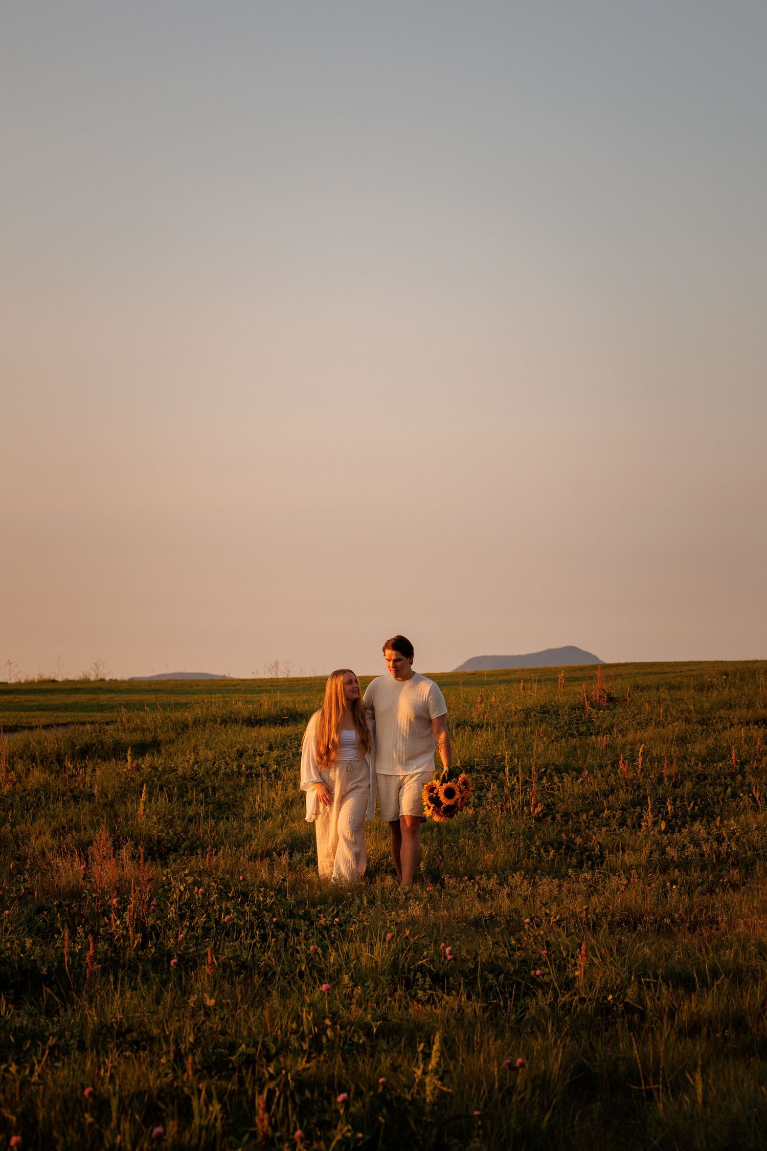 Two people walk together across a grassy hillside at sunset in a romantic golden hour setting.
