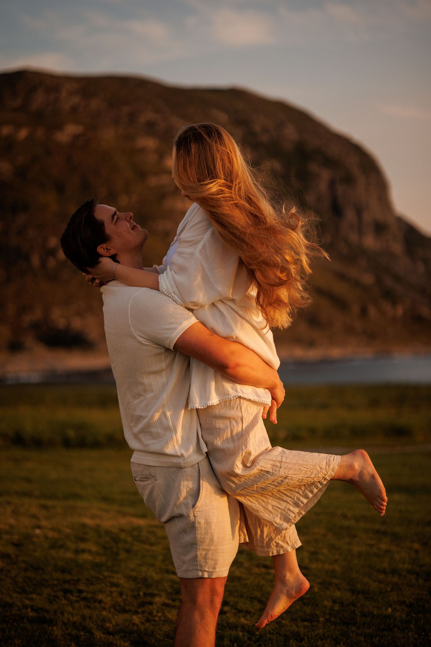 A couple in white outfits embrace and spin playfully in a golden-lit field at sunset with mountains in the background.