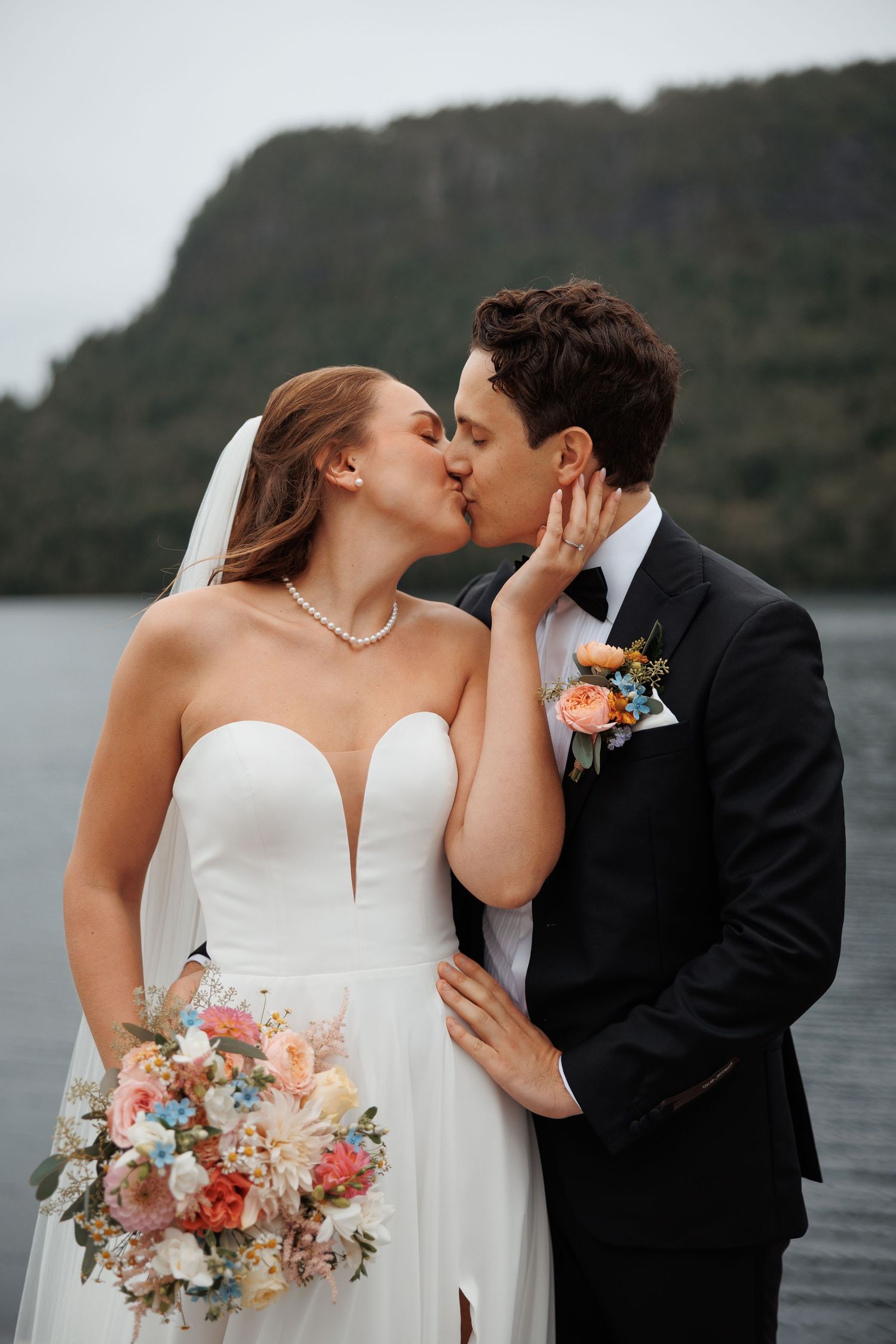 Romantic couple shares passionate kisses by a serene lake in a misty coastal landscape for their wedding photos.