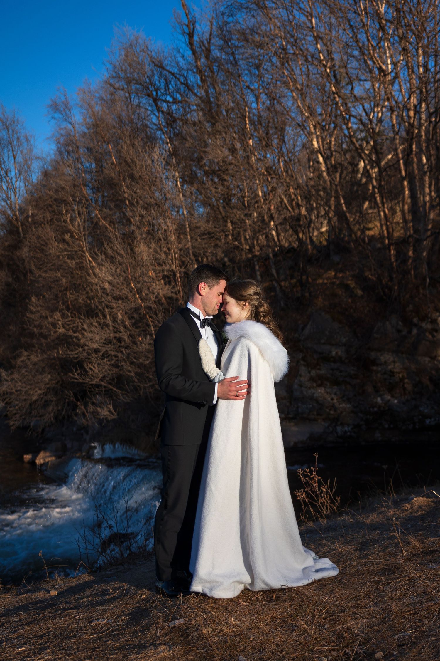 Elegant couple in winter wedding attire embrace against backdrop of bare trees and snow.