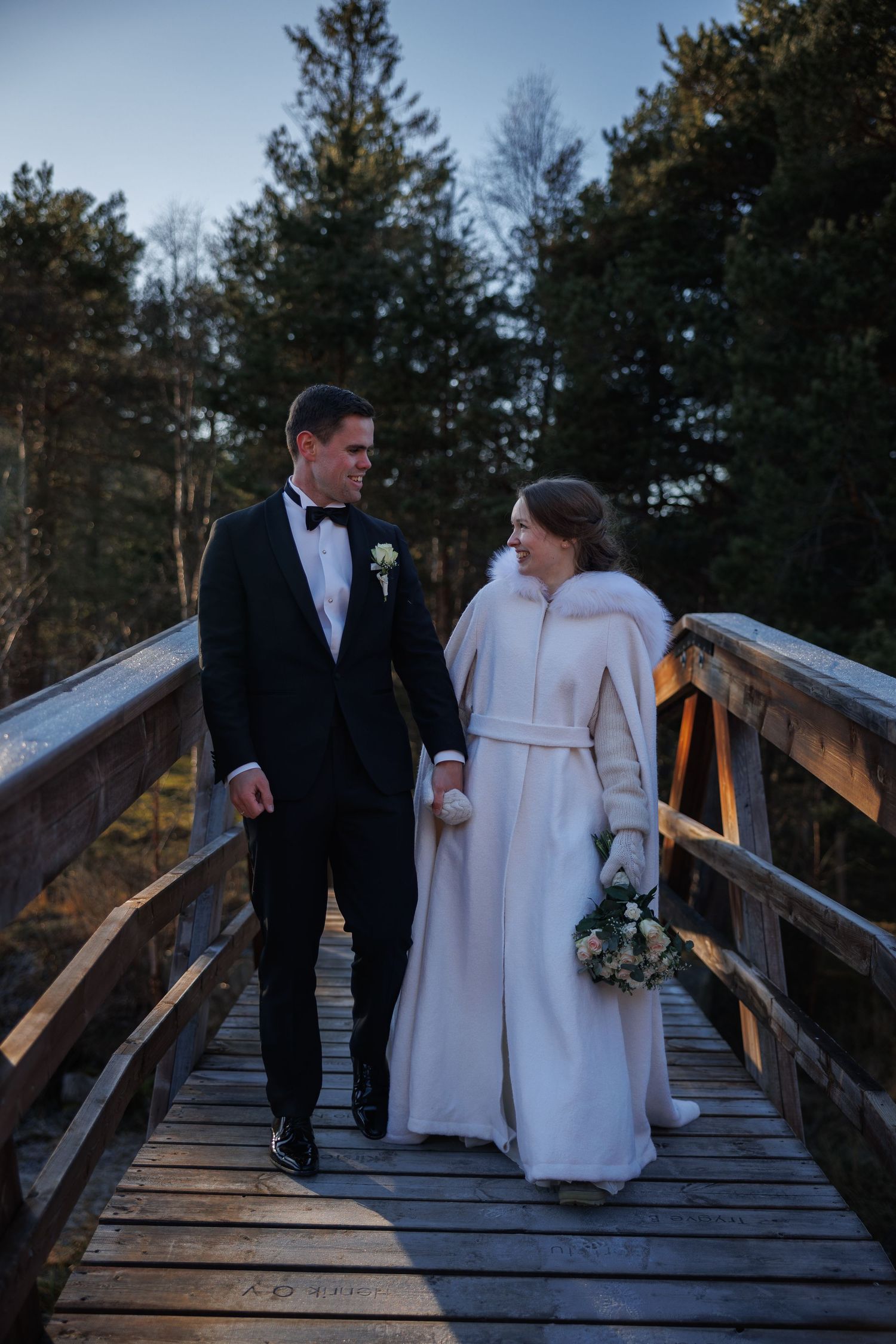 Bride and groom walking together on a wooden bridge in winter wedding attire.