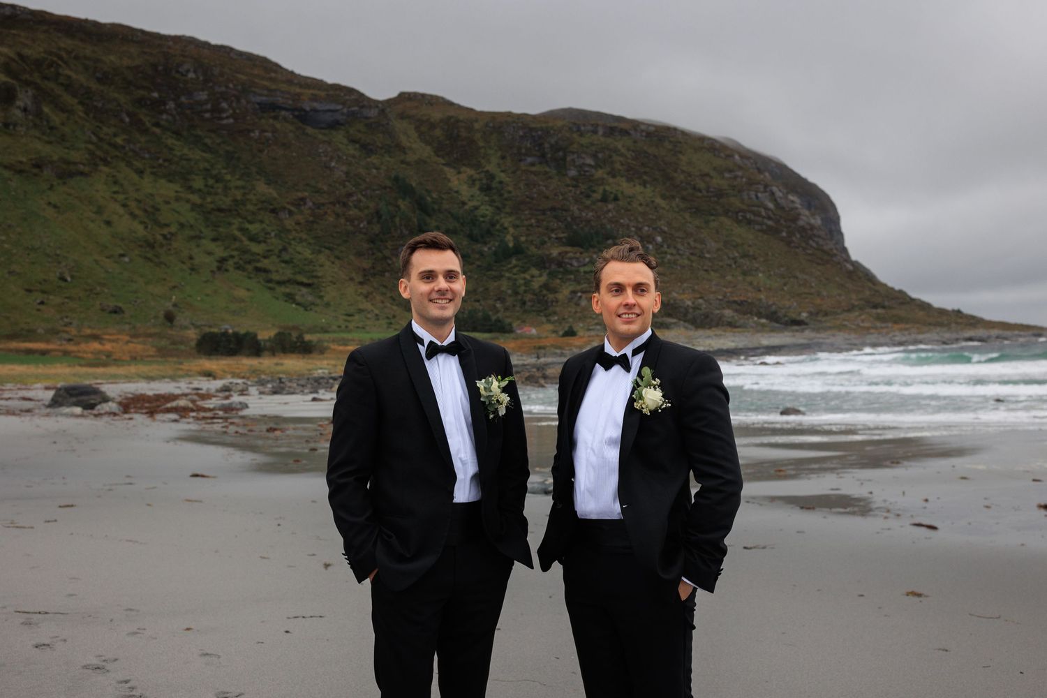 Two groomsmen in classic black tuxedos pose on beach with dramatic coastal cliffs behind them.