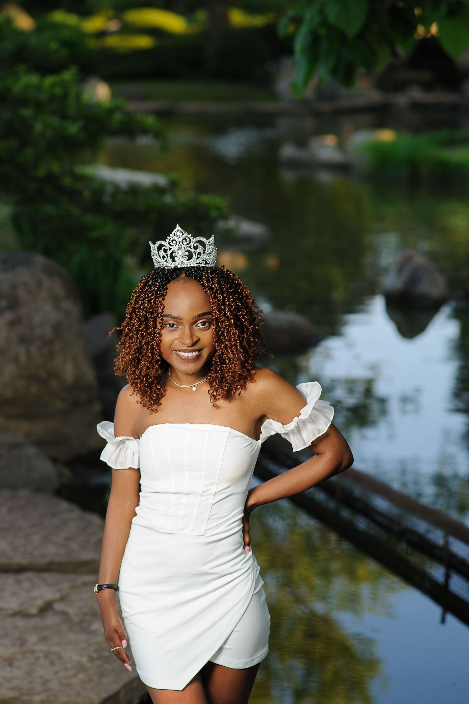A person in a white off-shoulder dress poses by a serene pond surrounded by lush greenery and reflective water.