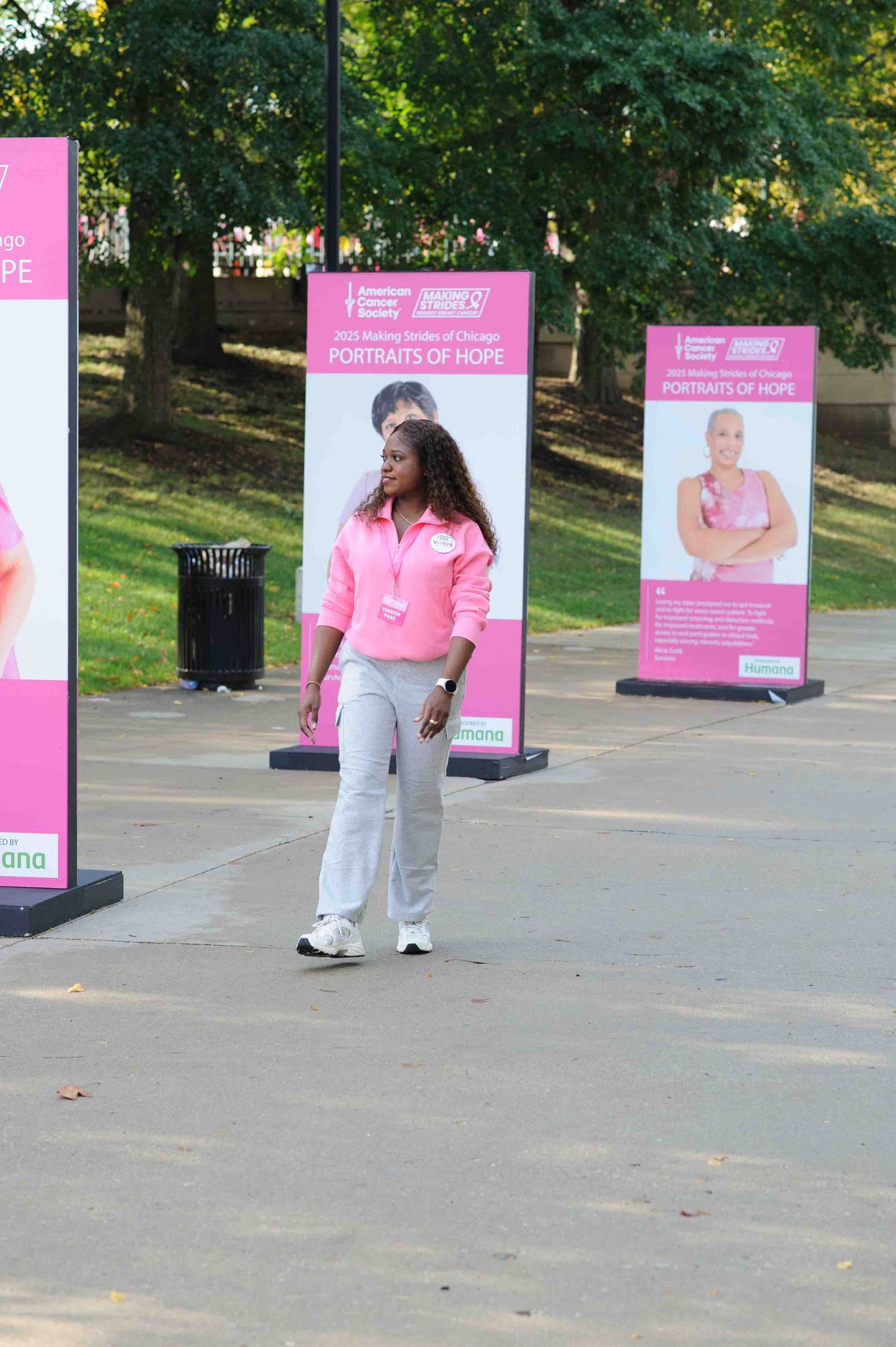 Pink advertising banners line a park pathway with people walking past on a sunny day. Tabeeka James.