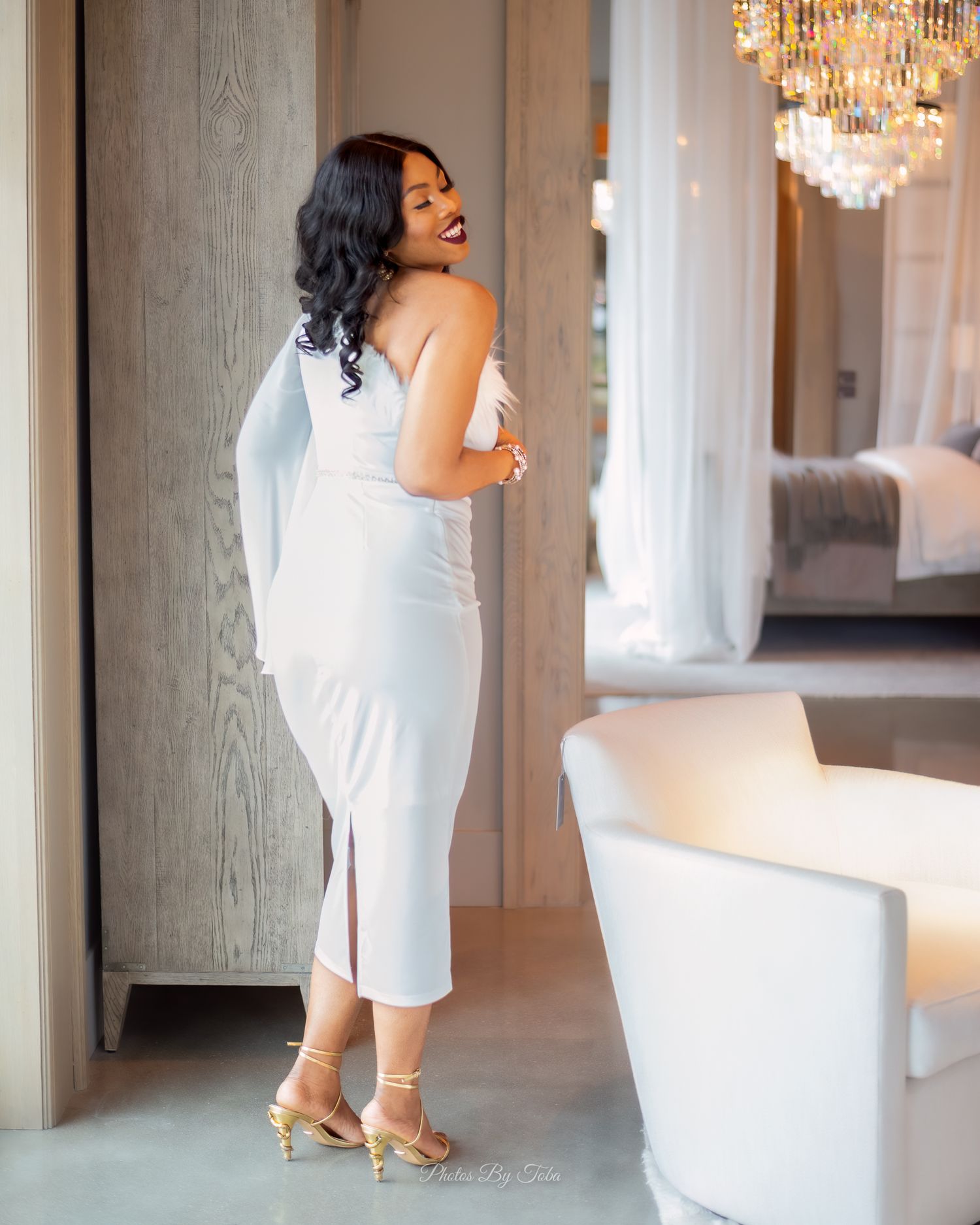 Woman in white midi dress and gold heels posing elegantly in a luxurious hotel room with chandelier.