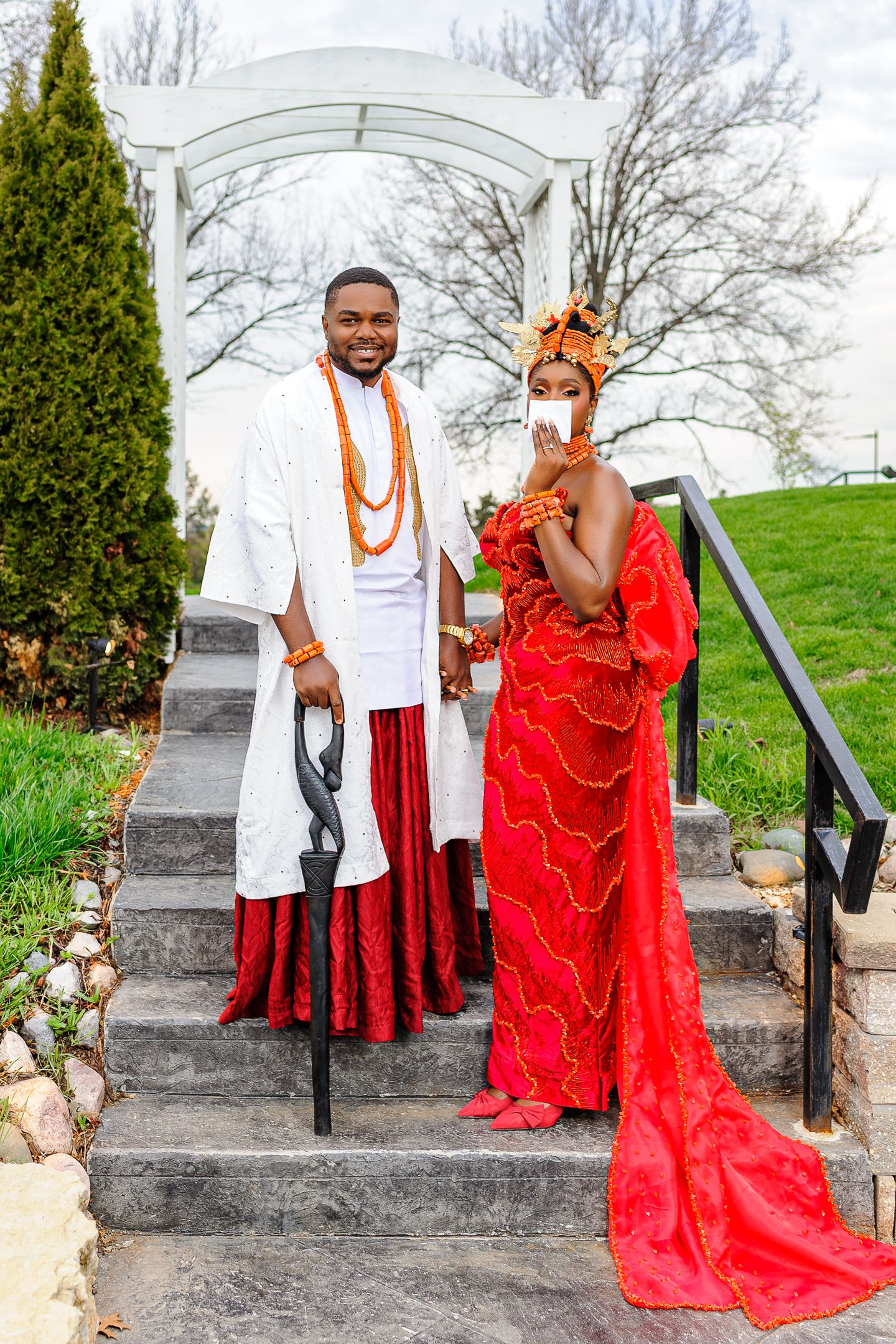 Couple in traditional Nigerian wedding attire, man in white and woman in red gown, posing on outdoor steps.