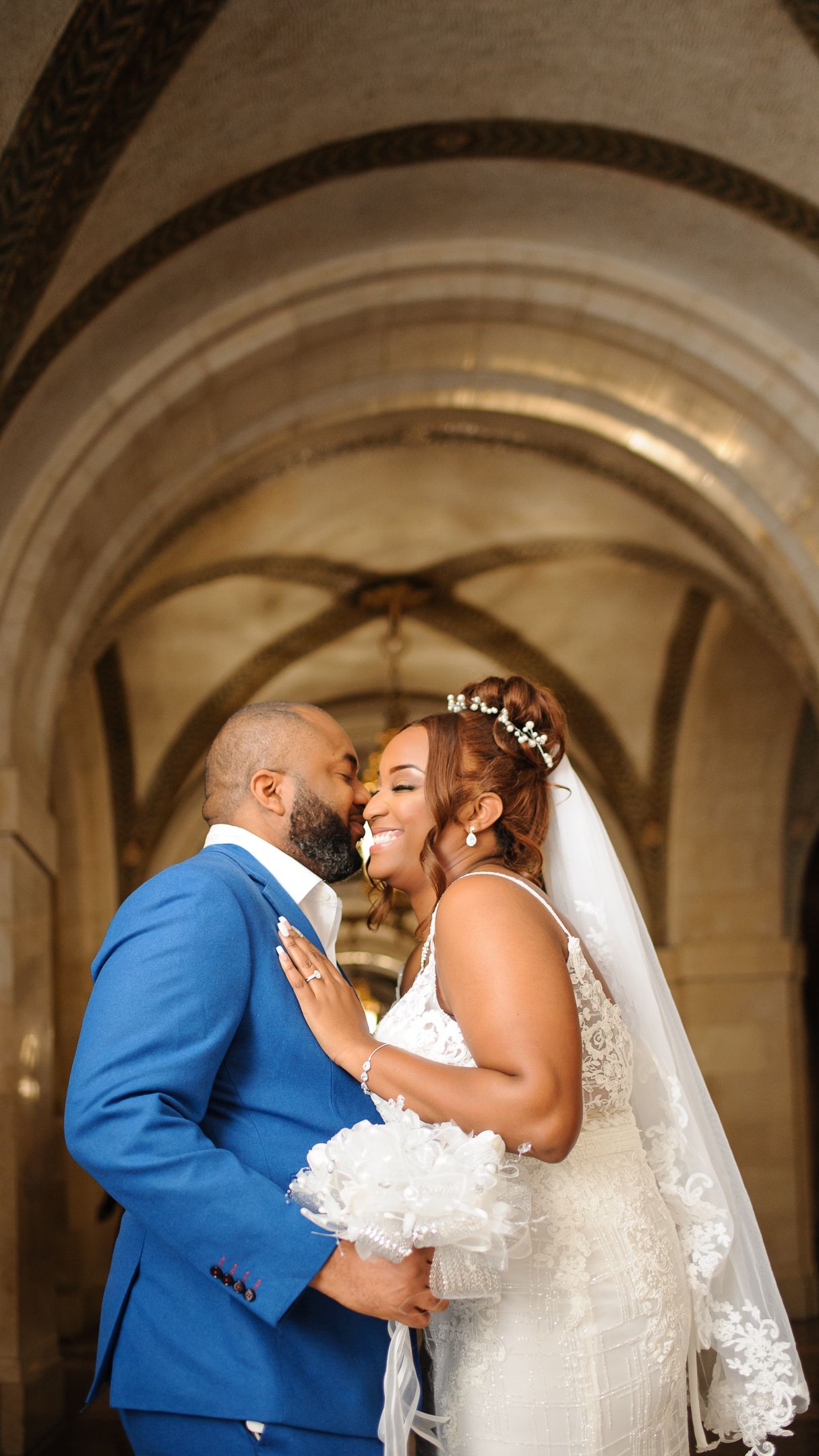A joyful bride in a white lace gown and groom in a blue suit share a romantic moment under stone arches.
