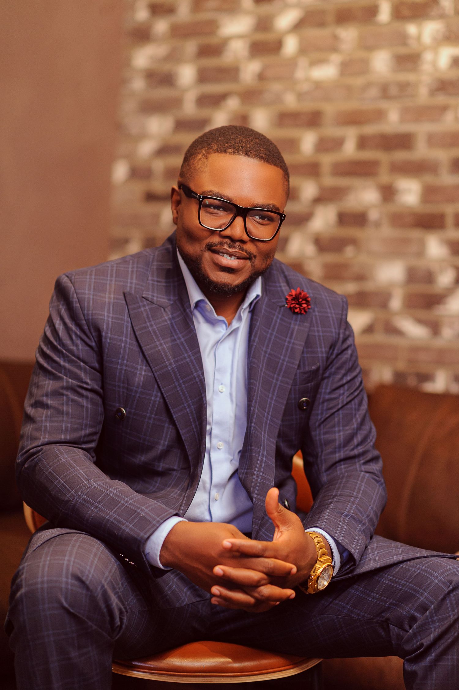 Man in plaid blue suit with glasses and red boutonniere sits confidently with hands clasped in stylish interior.