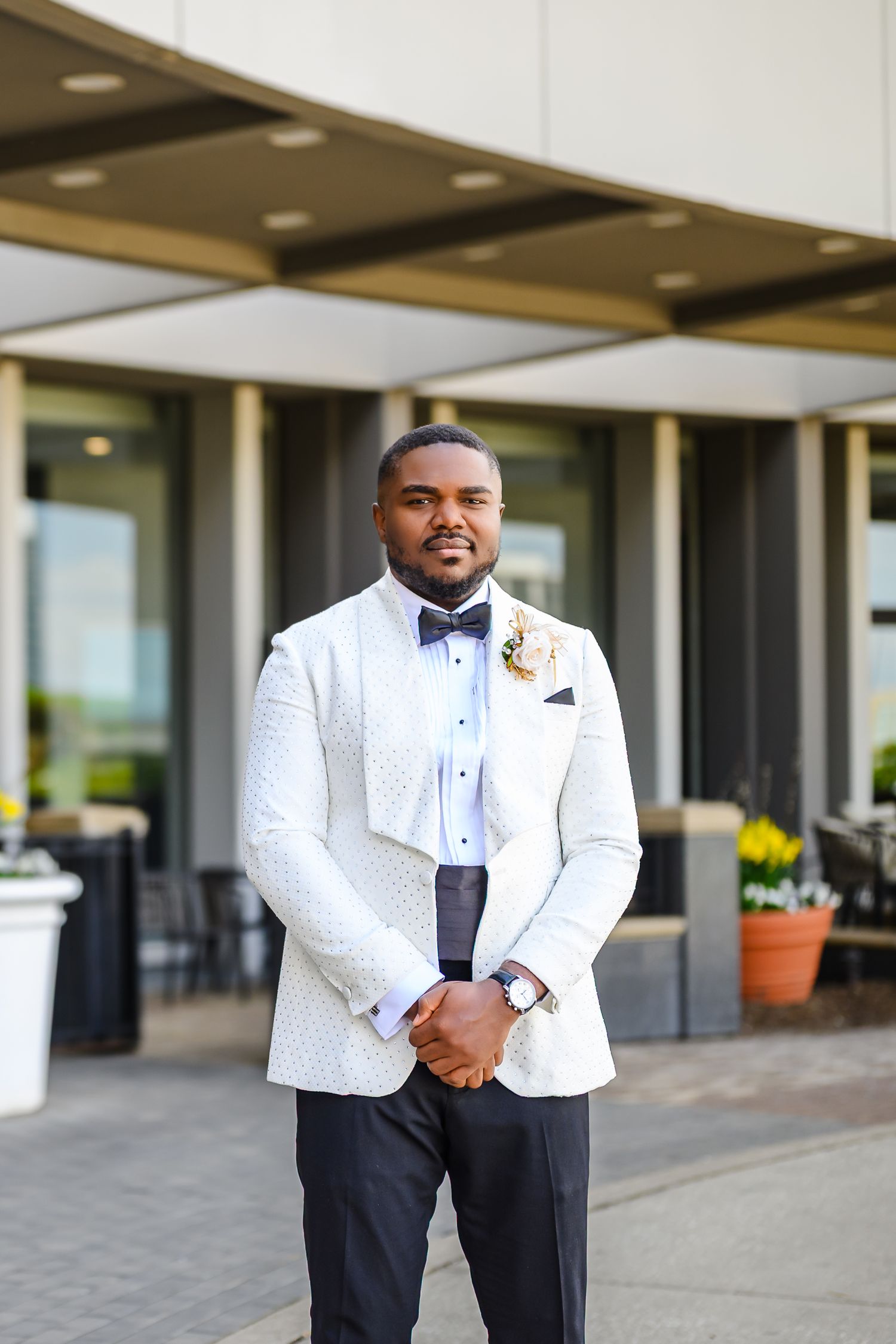 A well-dressed groom in a white tuxedo jacket with bow tie and boutonniere poses outside an elegant venue.