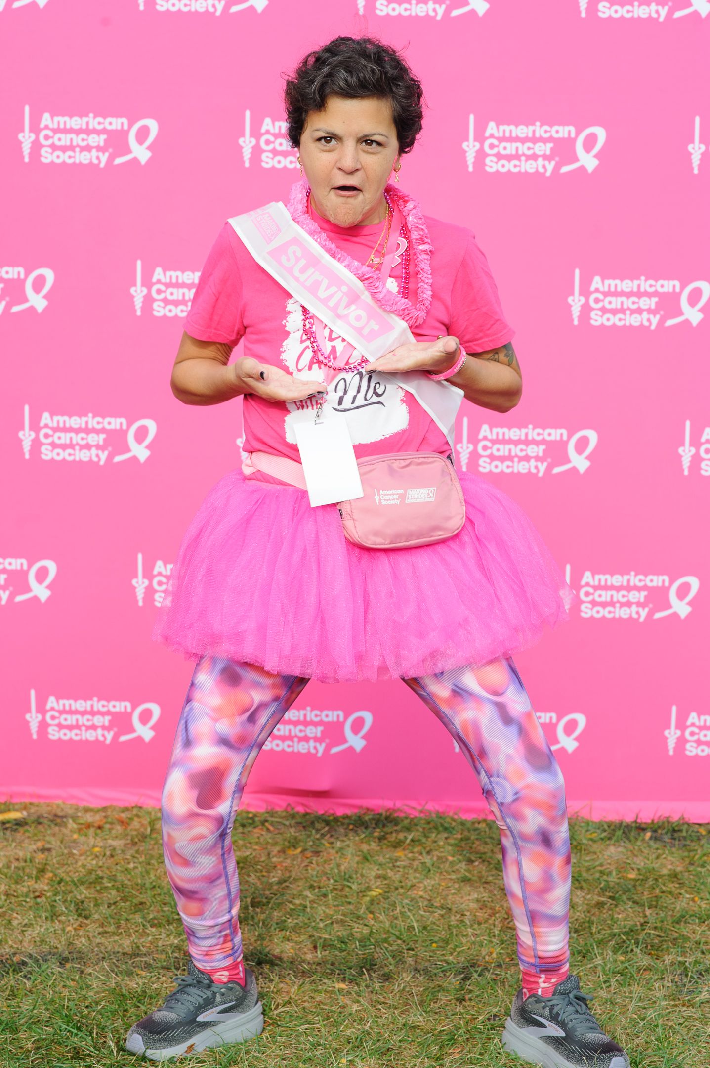 Person wearing pink tutu and tie-dye leggings poses playfully against pink charity event backdrop.