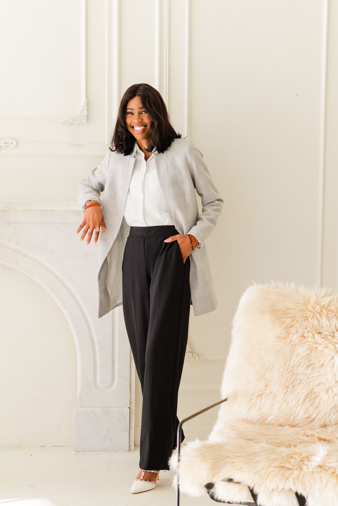 Smiling woman in gray coat, white blouse, and black trousers standing by a white fireplace in a bright room.