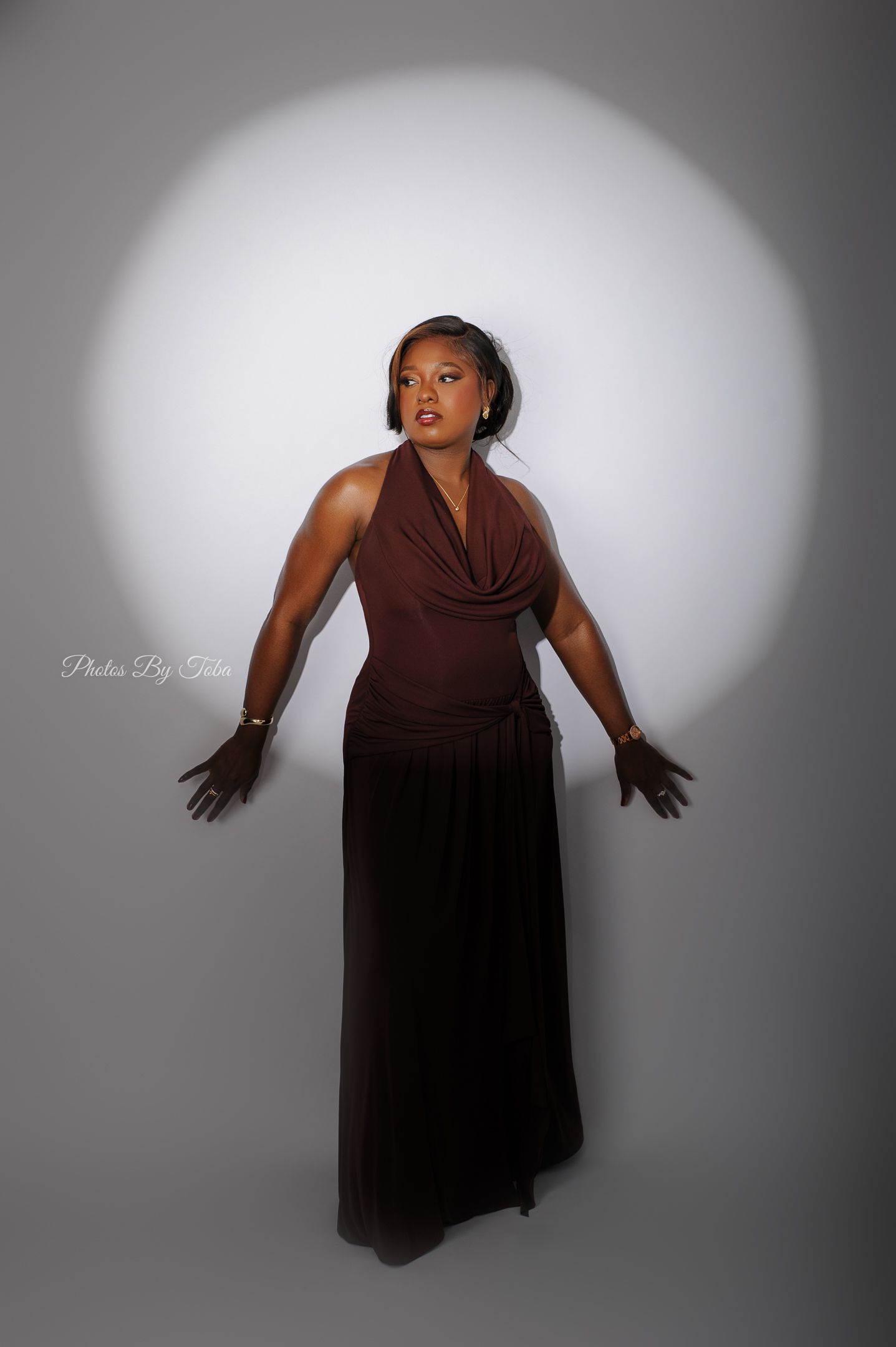 A woman in a brown halter maxi dress poses gracefully under a circular spotlight against a gray studio background.