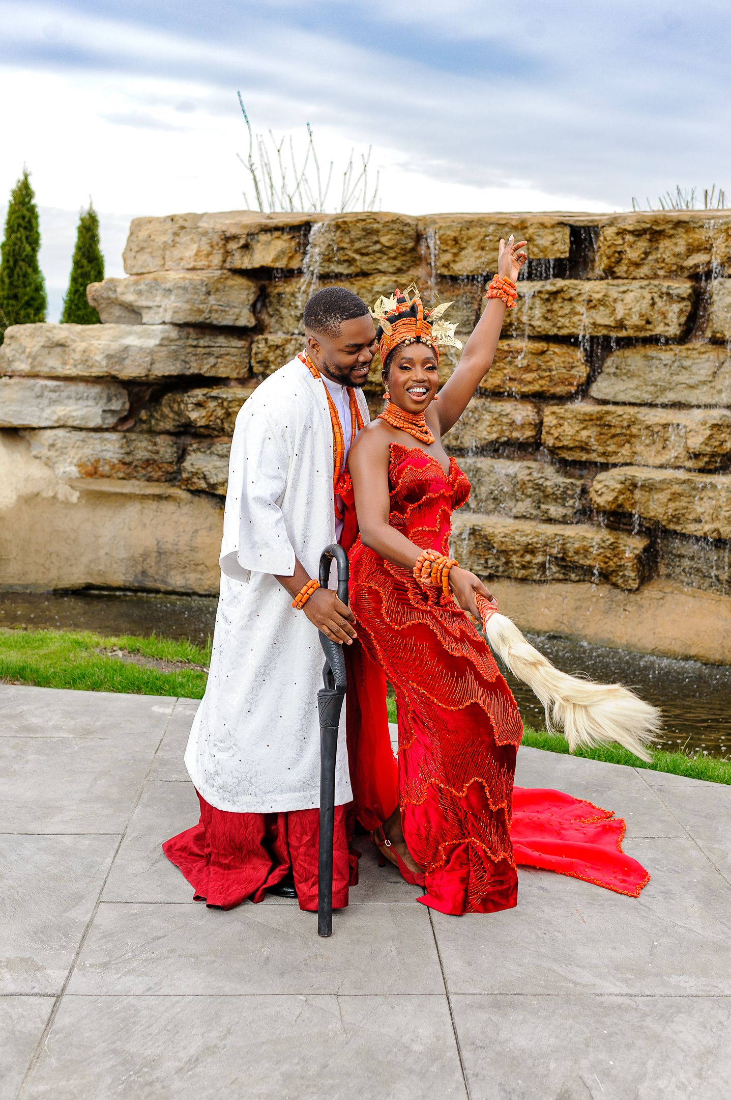 Nigerian wedding couple in traditional attire celebrate outdoors, bride raises hand joyfully.