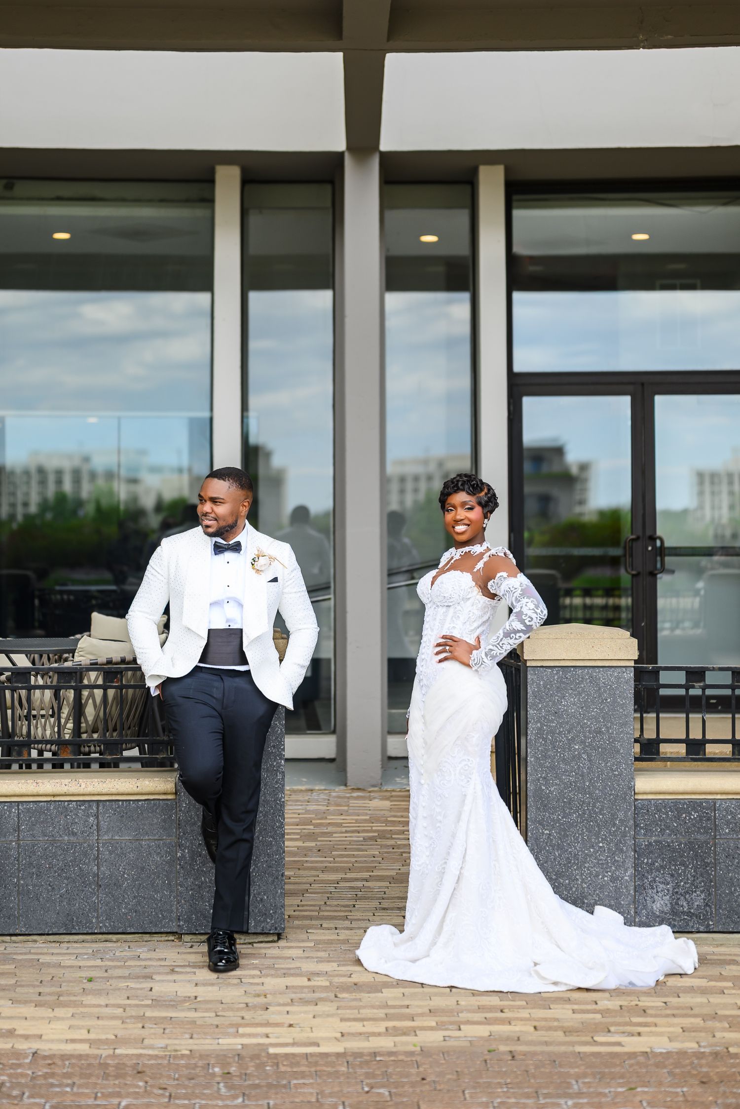 Elegant Black couple in wedding attire pose outside modern venue, bride in white gown, groom in white blazer.