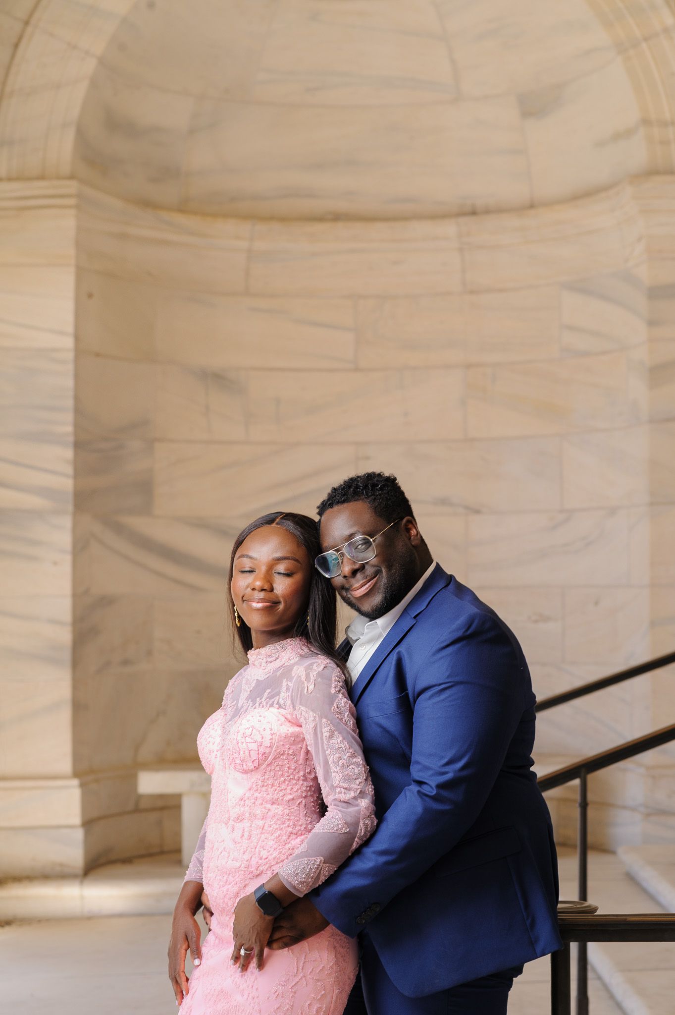 Couple posing elegantly in a stone archway, woman in pink lace gown and man in navy blue suit with sunglasses.