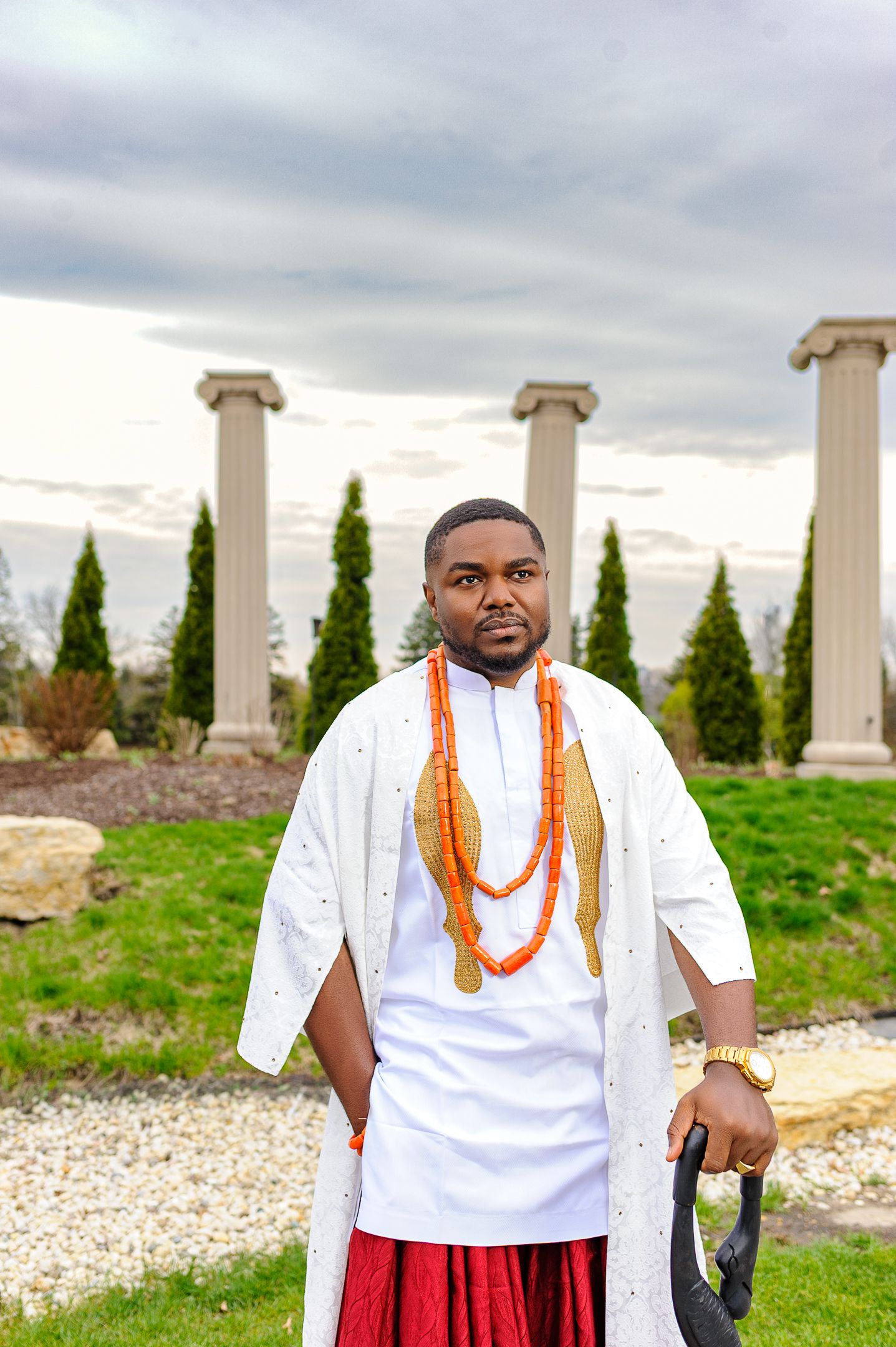 Nigerian groom in white agbada with coral beads and red wrapper holding ceremonial staff at outdoor venue.