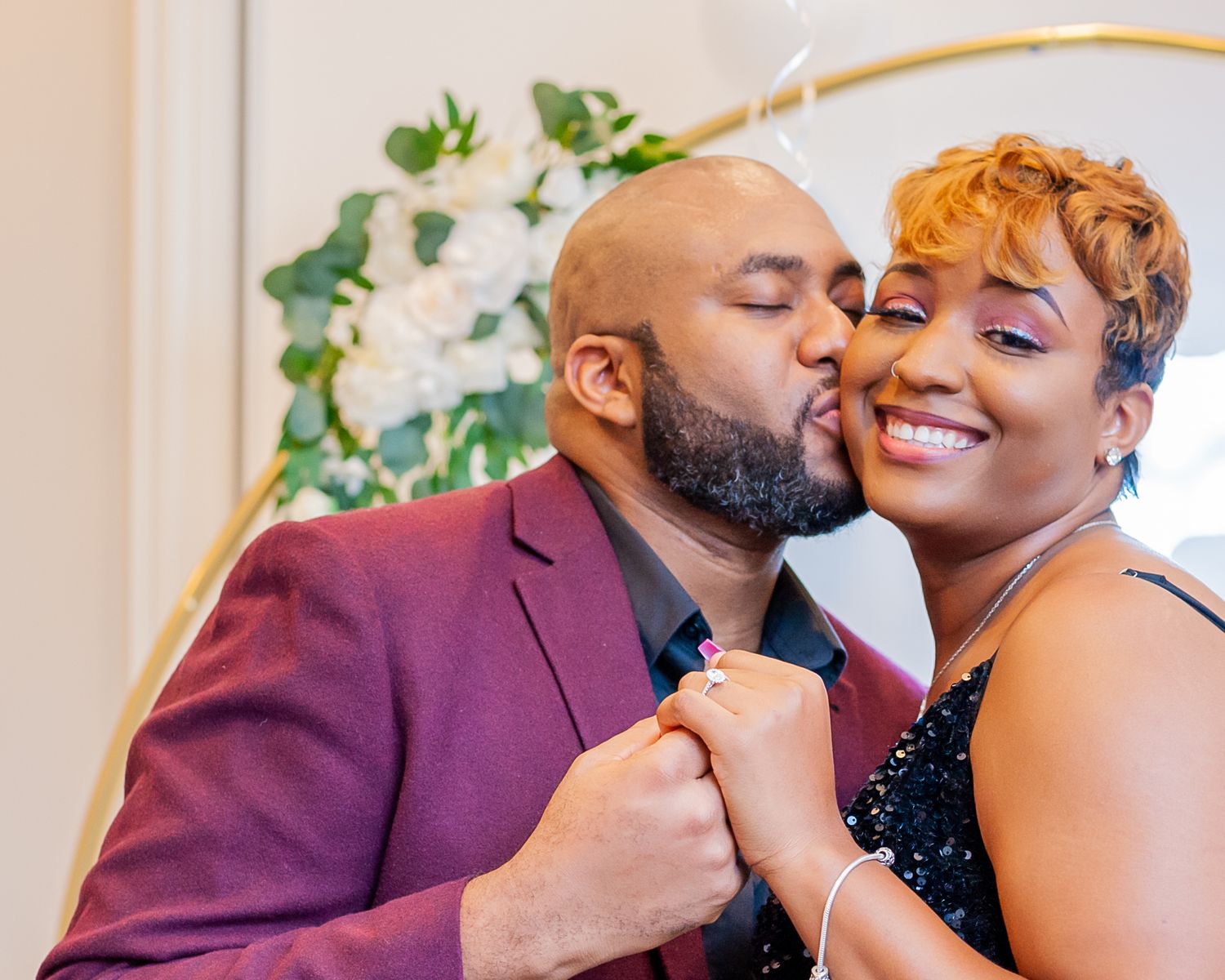 A man in a burgundy suit kisses a smiling woman on the cheek at a romantic wedding ceremony.