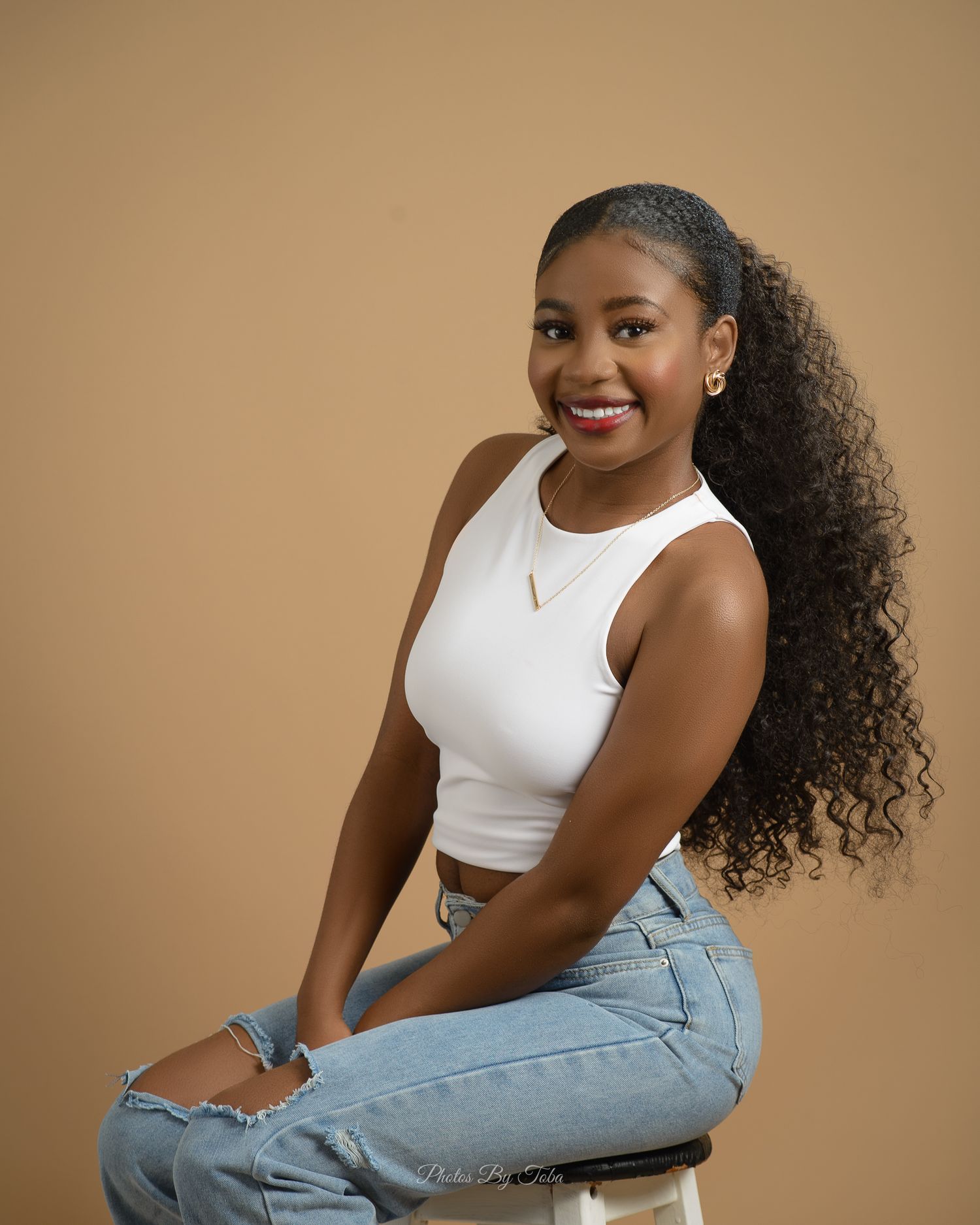 A casual fashion portrait with someone wearing a white crop top and jeans against a tan background.