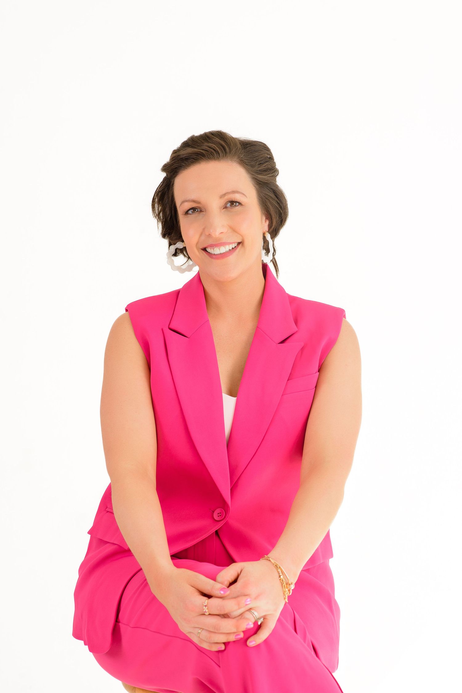 Professional wearing a vibrant hot pink sleeveless blazer sits casually while smiling at the camera.