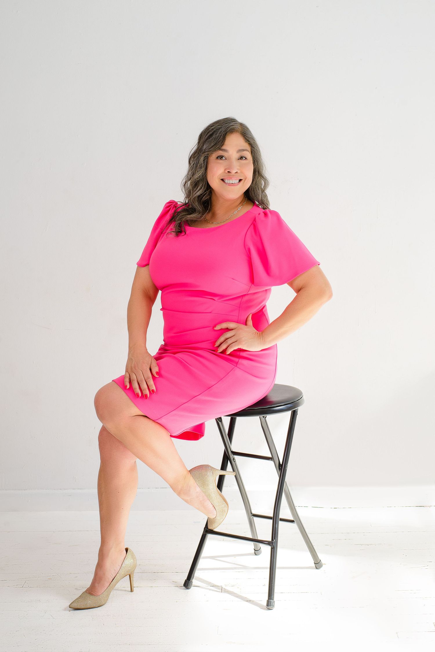 A person in a vibrant hot pink dress poses confidently on a black stool against a white studio backdrop.