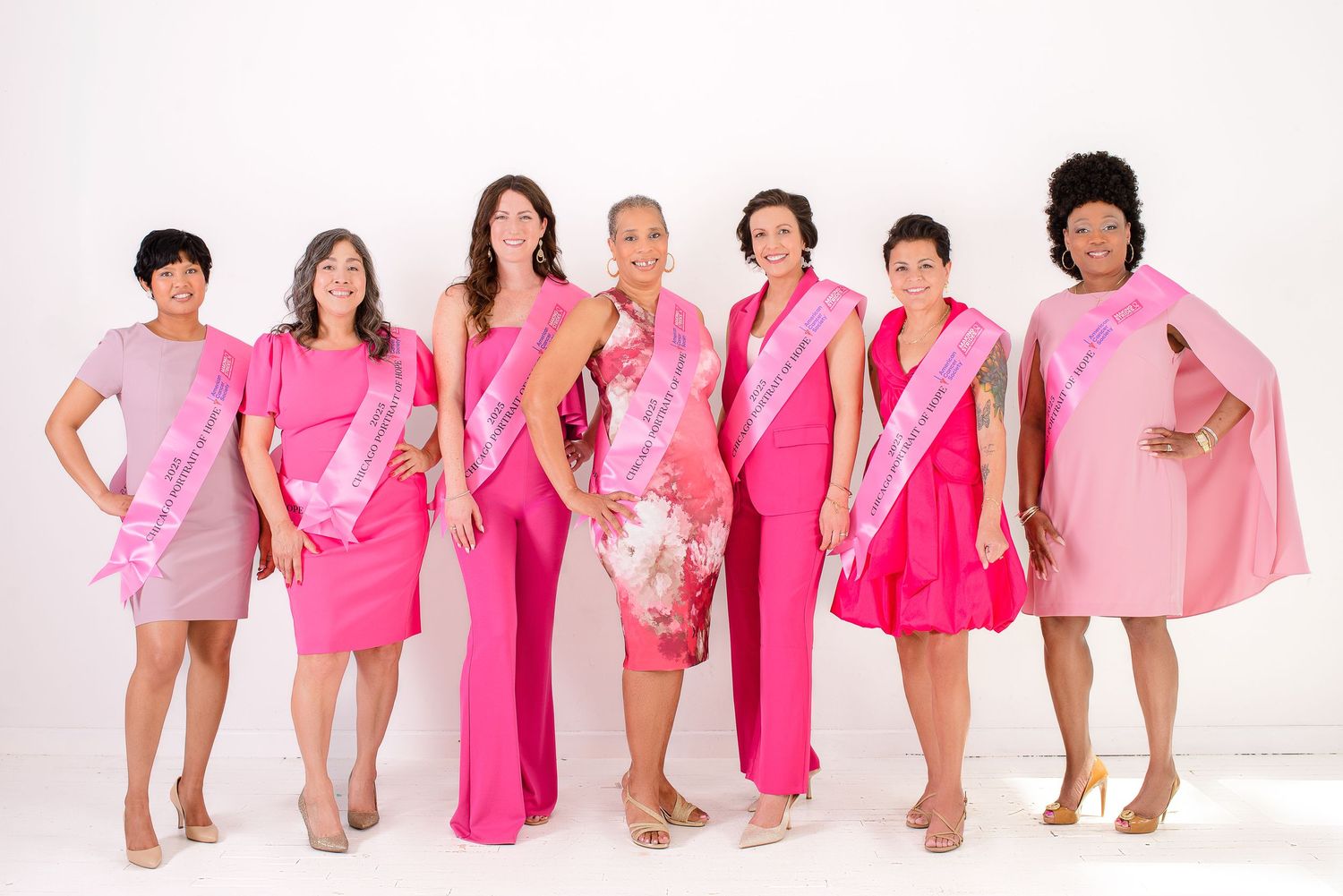 A group of people wearing various shades of pink clothing pose together in a white studio setting.