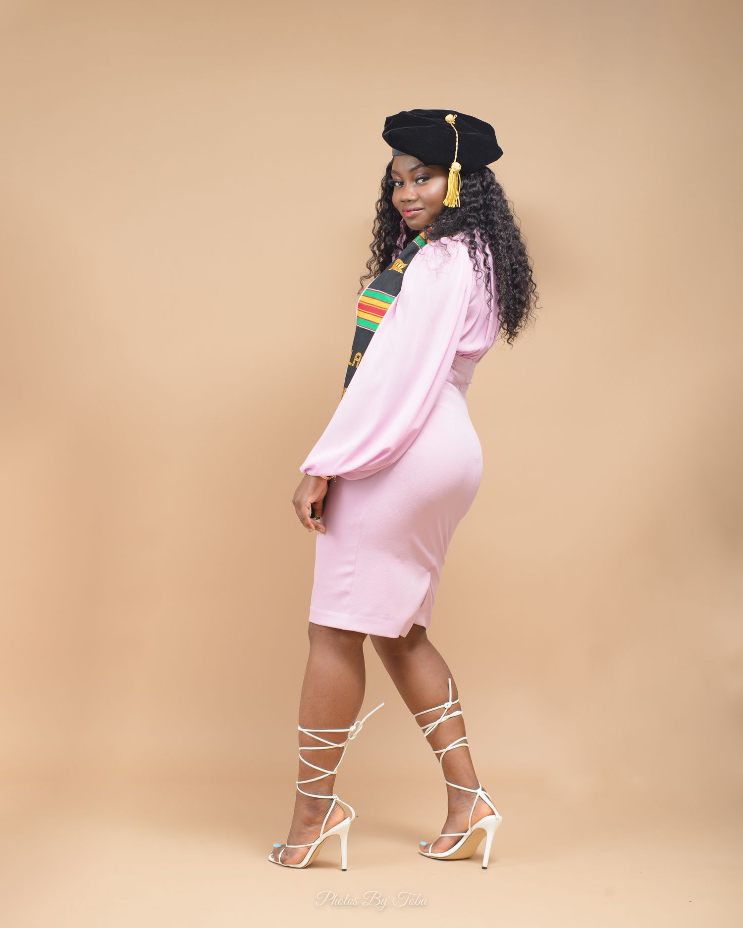 Graduation photo shoot featuring a pink dress and strappy heels against a beige studio background.