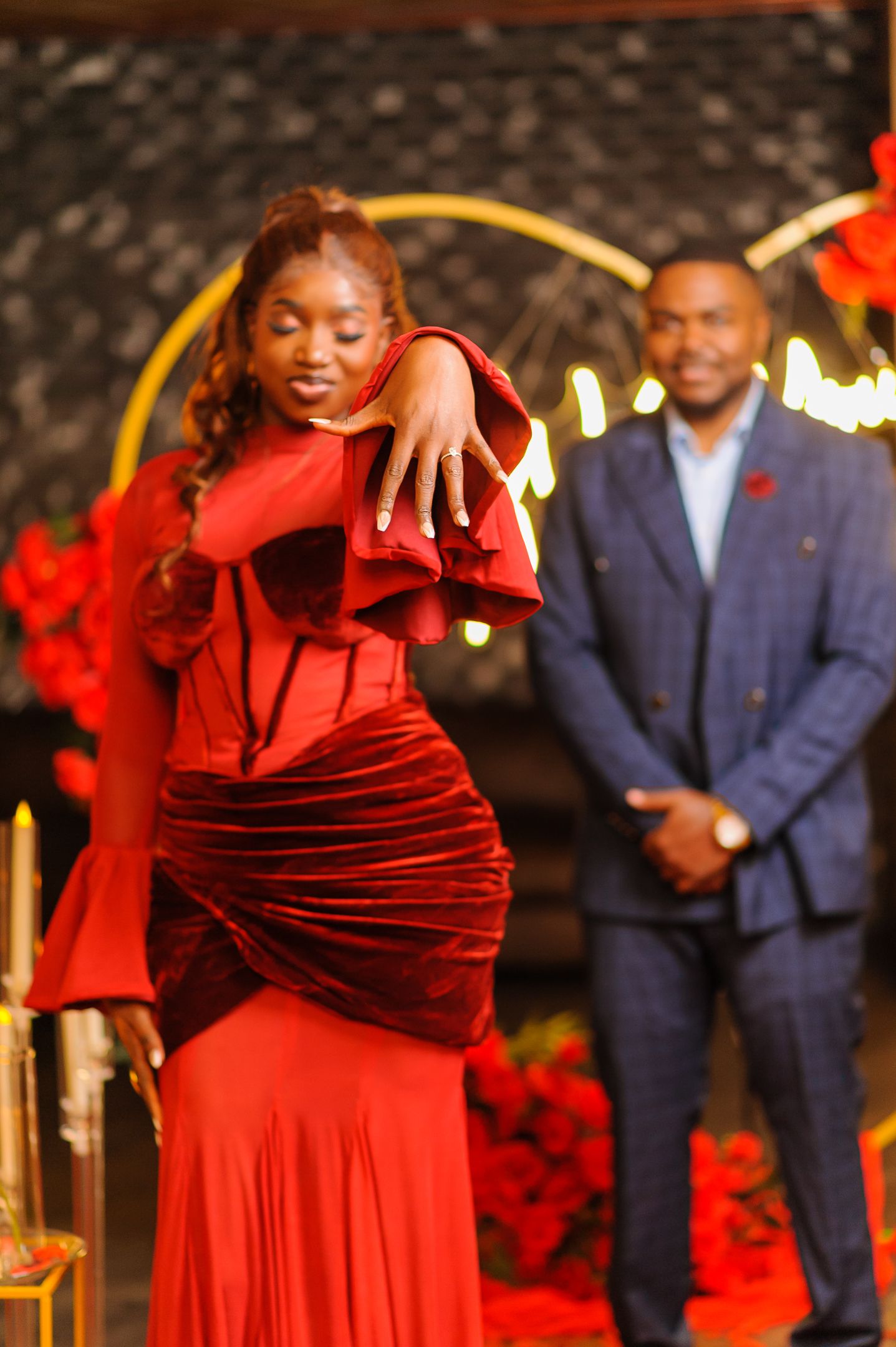 Woman in elegant red velvet gown posing at a festive event with a man in blue suit smiling in background.