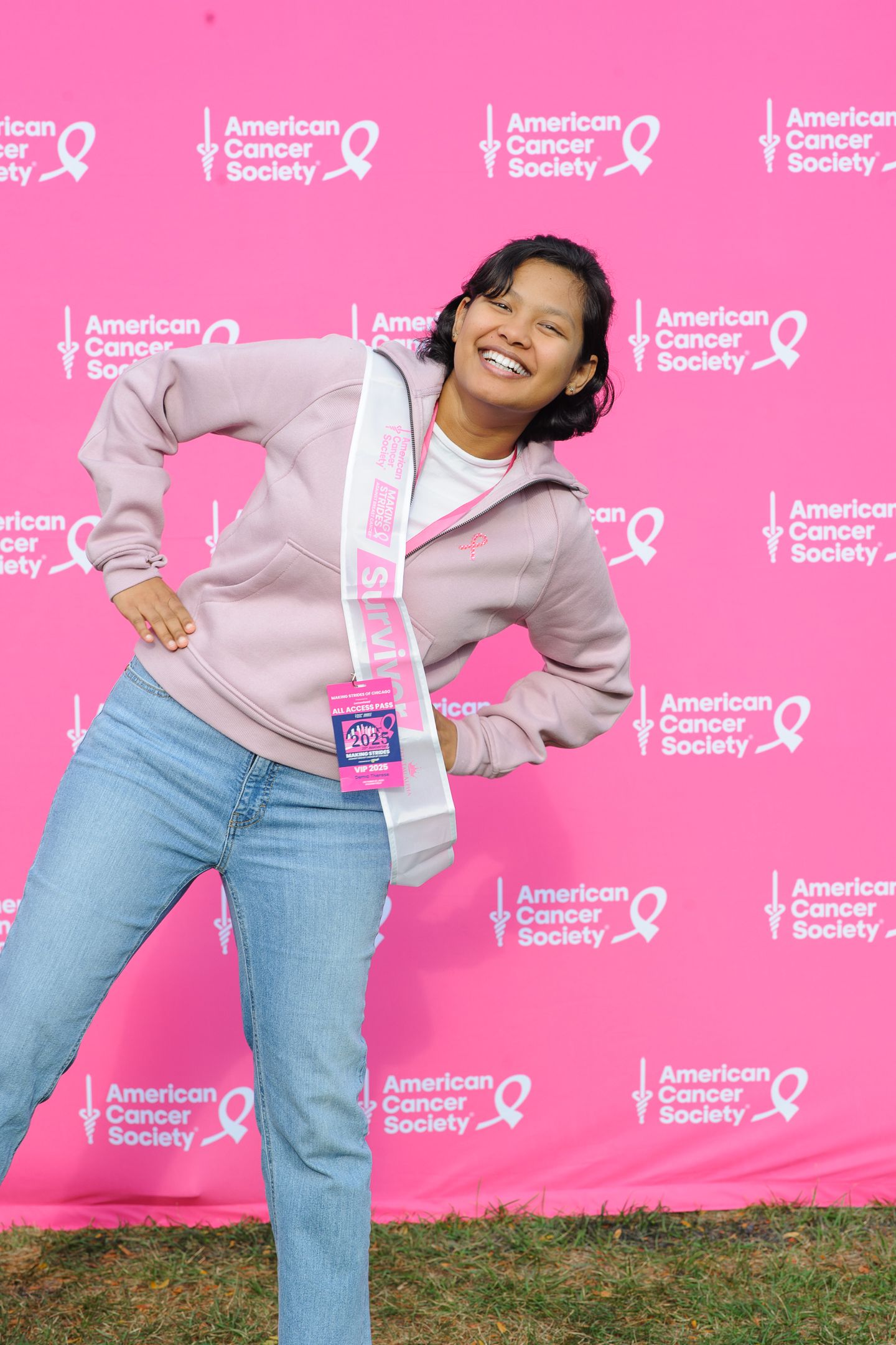 Person in grey sweater and jeans poses energetically against pink American Cancer Society backdrop.