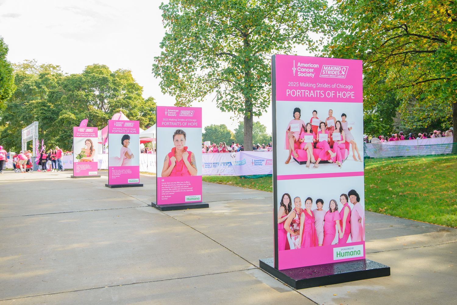 Pink promotional banners displaying K-pop group advertisements line an outdoor pathway in a park setting.