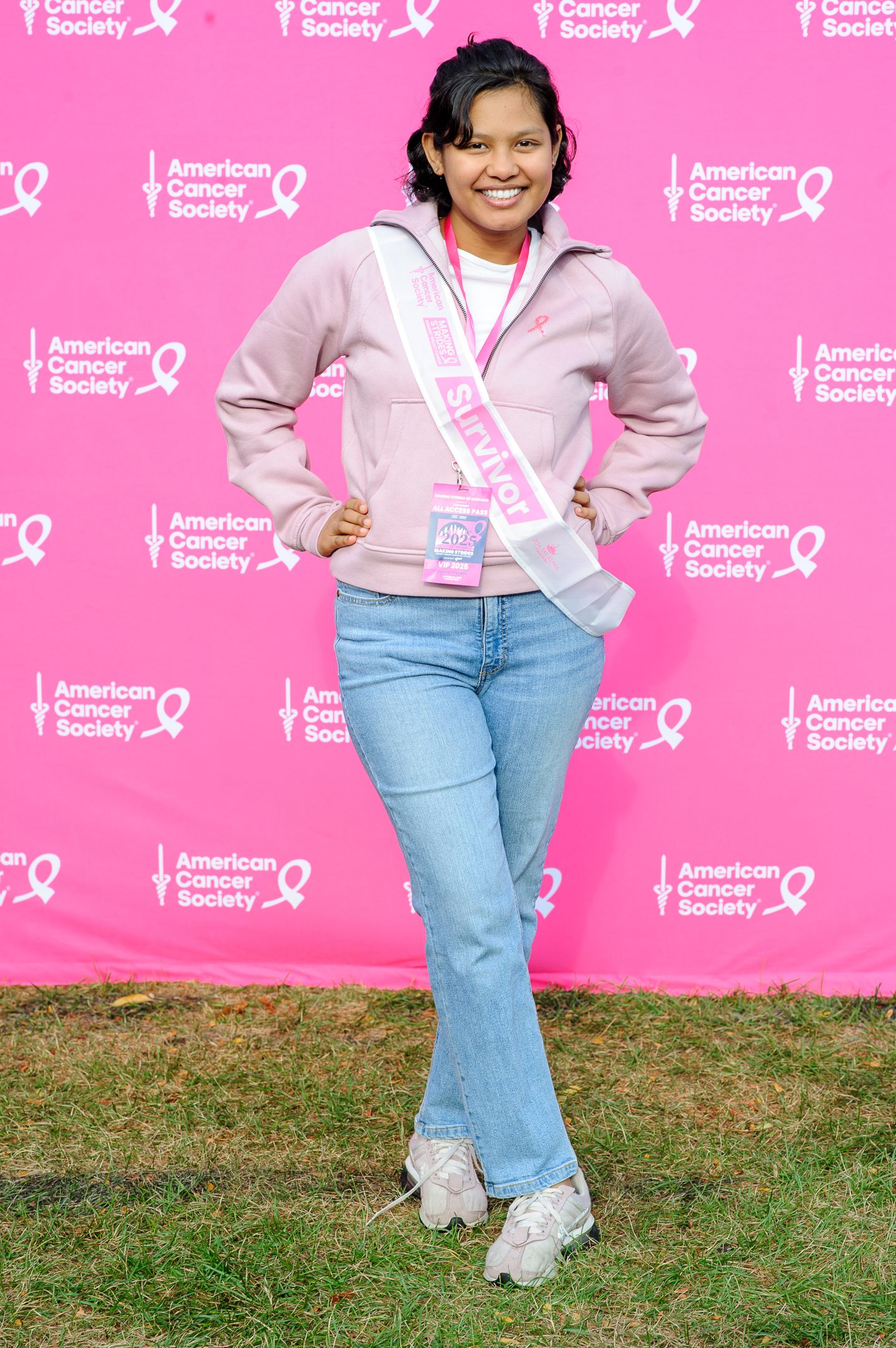 Woman in light pink jacket and jeans poses cheerfully against pink backdrop at charity event.