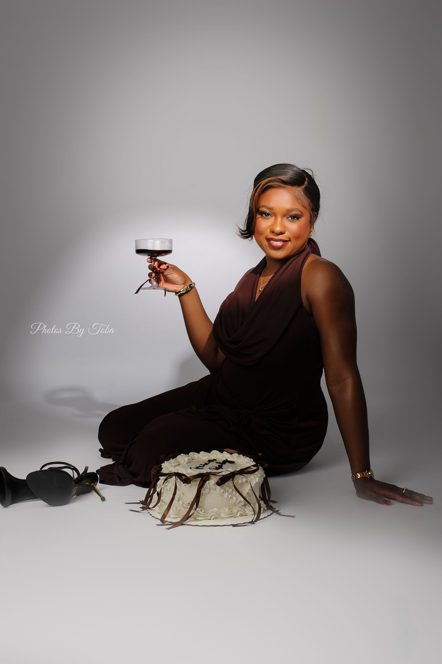 A smiling woman in a brown jumpsuit sits on a white studio floor, holding a wine glass, with a handbag beside her.