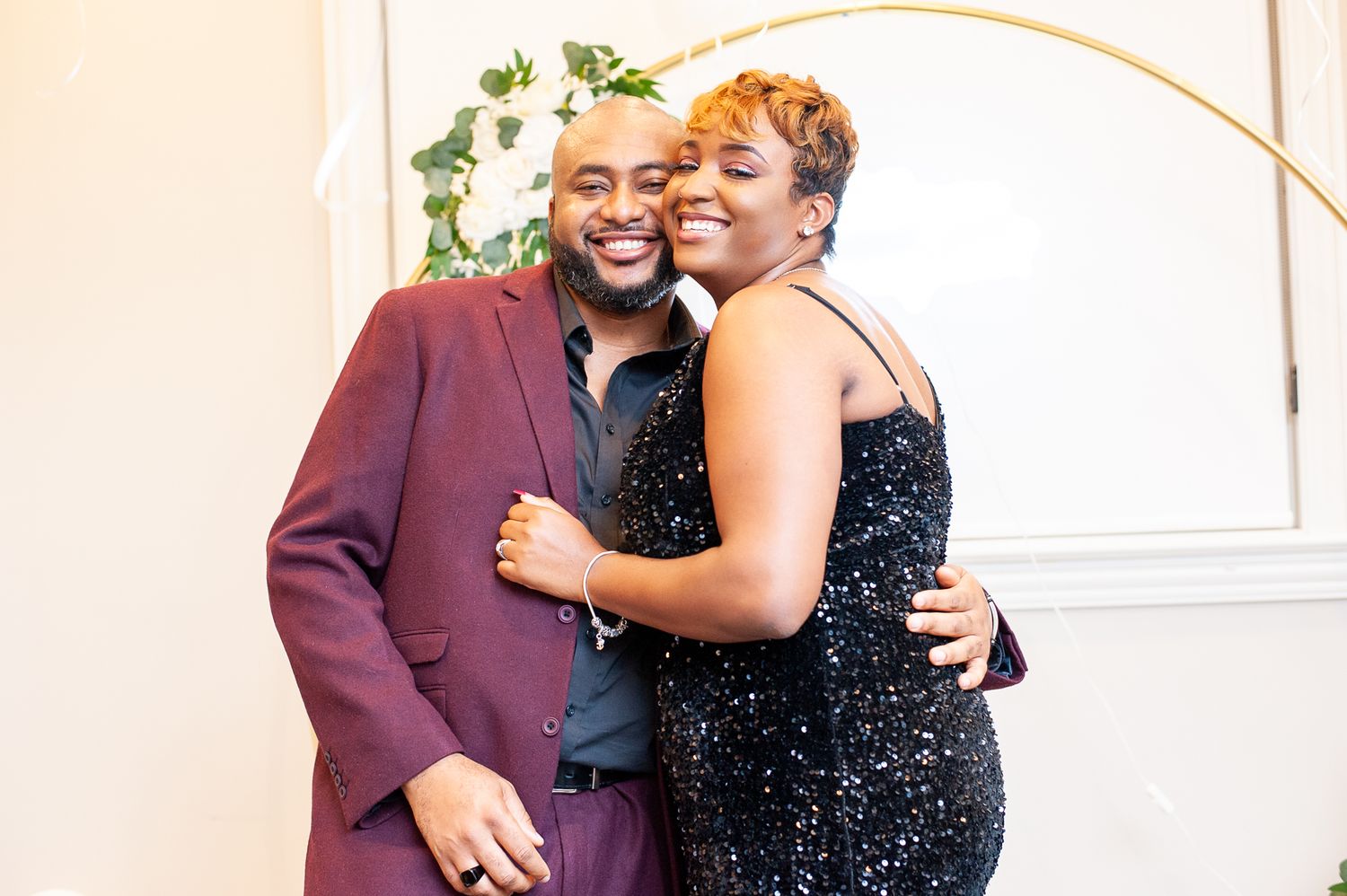 Couple embracing and smiling at a formal event, wearing elegant black sequin dress and burgundy suit.