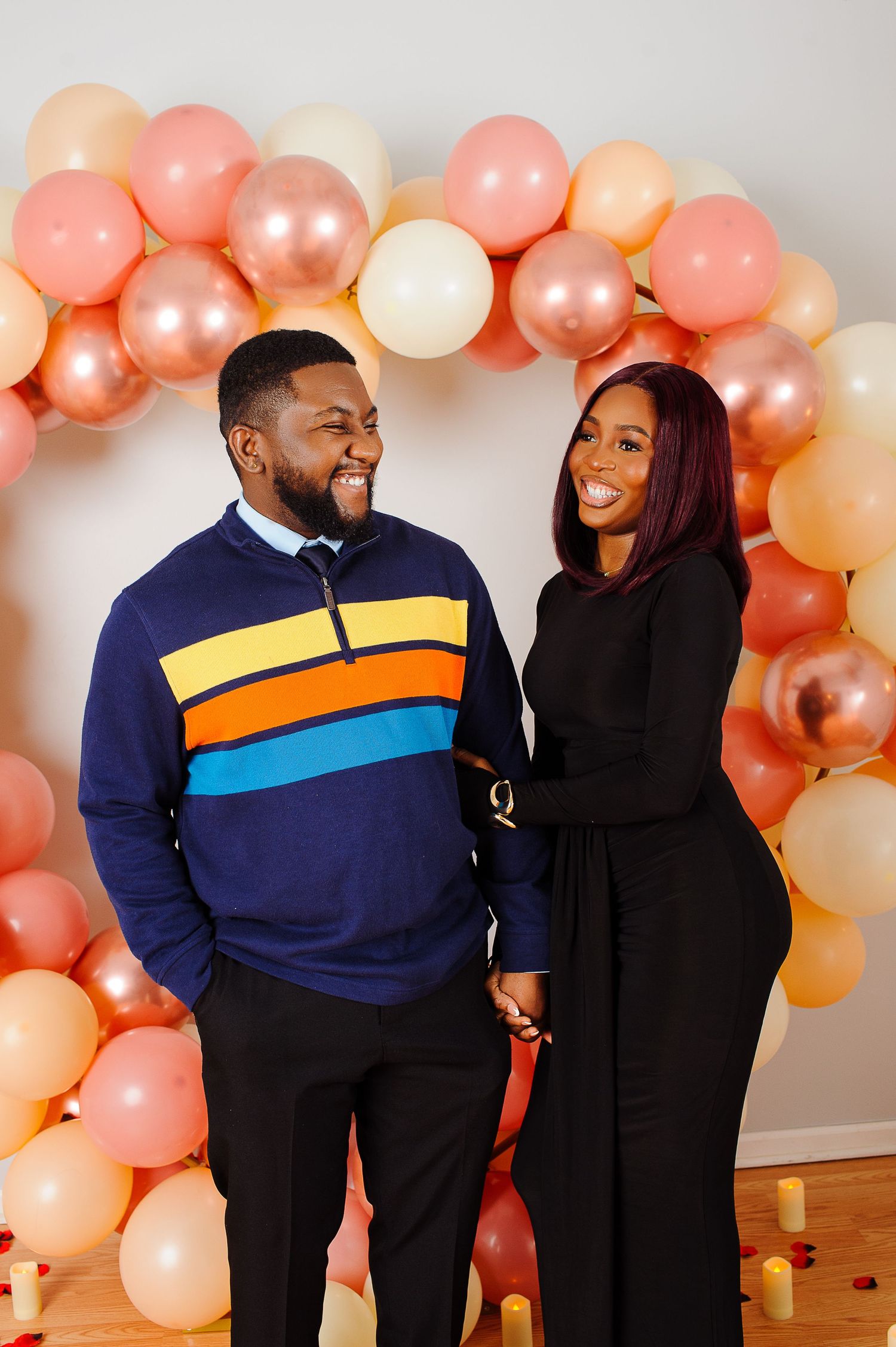 Happy couple posing together in front of a colorful balloon arch backdrop, smiling and holding hands.