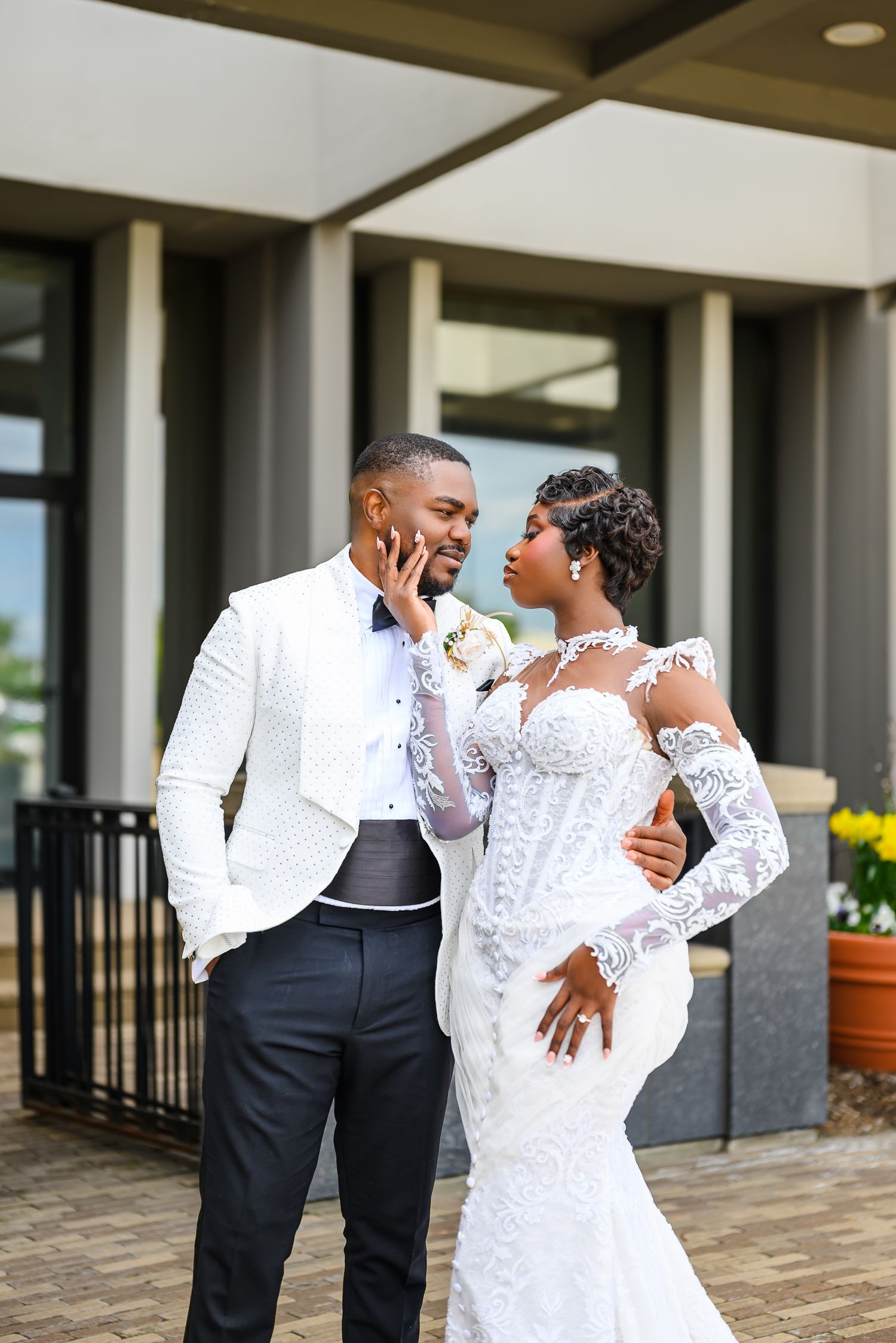 A bride and groom share a romantic moment outdoors, she in lace gown, he in white jacket and black trousers.